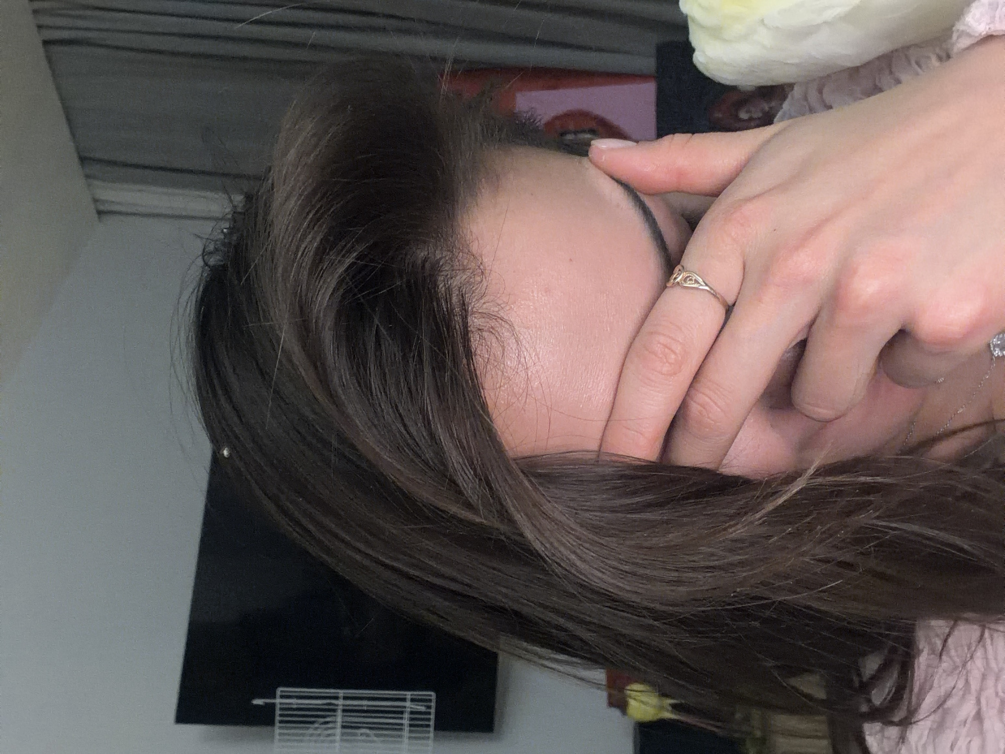 An indoor, close-up, and vertically oriented shot depicts a person, likely a woman, with dark brown hair covering the right side of her face with her right hand. Her forehead and the top part of her face are visible between her hair and hand. Her eyes are entirely covered by her fingers and palm.
The person's long, dark brown hair cascades over her forehead and the side of her face, with some lighter strands visible, particularly around the hairline where finer baby hairs are apparent. Her skin appears fair.
On her right hand, a golden ring with an intricate, possibly Celtic knot-like design, is worn on her ring finger. A second, silver-colored ring, possibly with a small clear stone, is partially visible on her pinky finger. A delicate silver-toned necklace chain is also visible around her neck. Her nails are short and neatly kept.
In the background, to the upper right, a light-colored, fluffy object, possibly a white bird or a decorative item, is partially visible. The person appears to be wearing a light-pink, textured garment, possibly a ruffled sleeve or top, on the right side of the frame. The lower-left background shows a dark, rectangular object, possibly a television or monitor screen, with a white, grid-like structure (perhaps a part of a cage or storage unit) in front of it. The overall setting appears to be a personal, indoor space.
The activity depicted is the person covering their face, which could suggest shyness, contemplation, tiredness, or shielding eyes from light. No text is visible in the image. The location context provided is Krasnoyarsk, Russia, although no specific visual cues in the image confirm this geographical detail.
