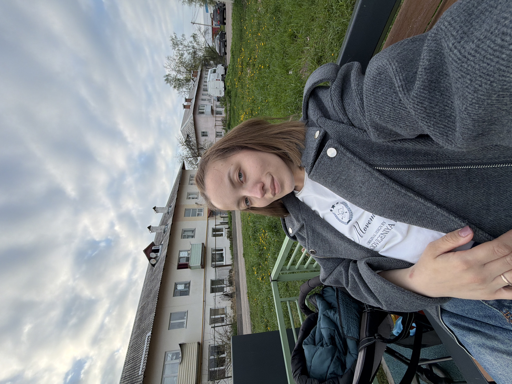 A woman is sitting on a bench in Reshetnikovo, Russia. She is wearing a gray jacket and a white t-shirt, and she has shoulder-length brown hair. Behind her is a multi-story residential building. There is a green lawn in the background with some yellow flowers, and several parked cars are visible further away. The sky is cloudy.