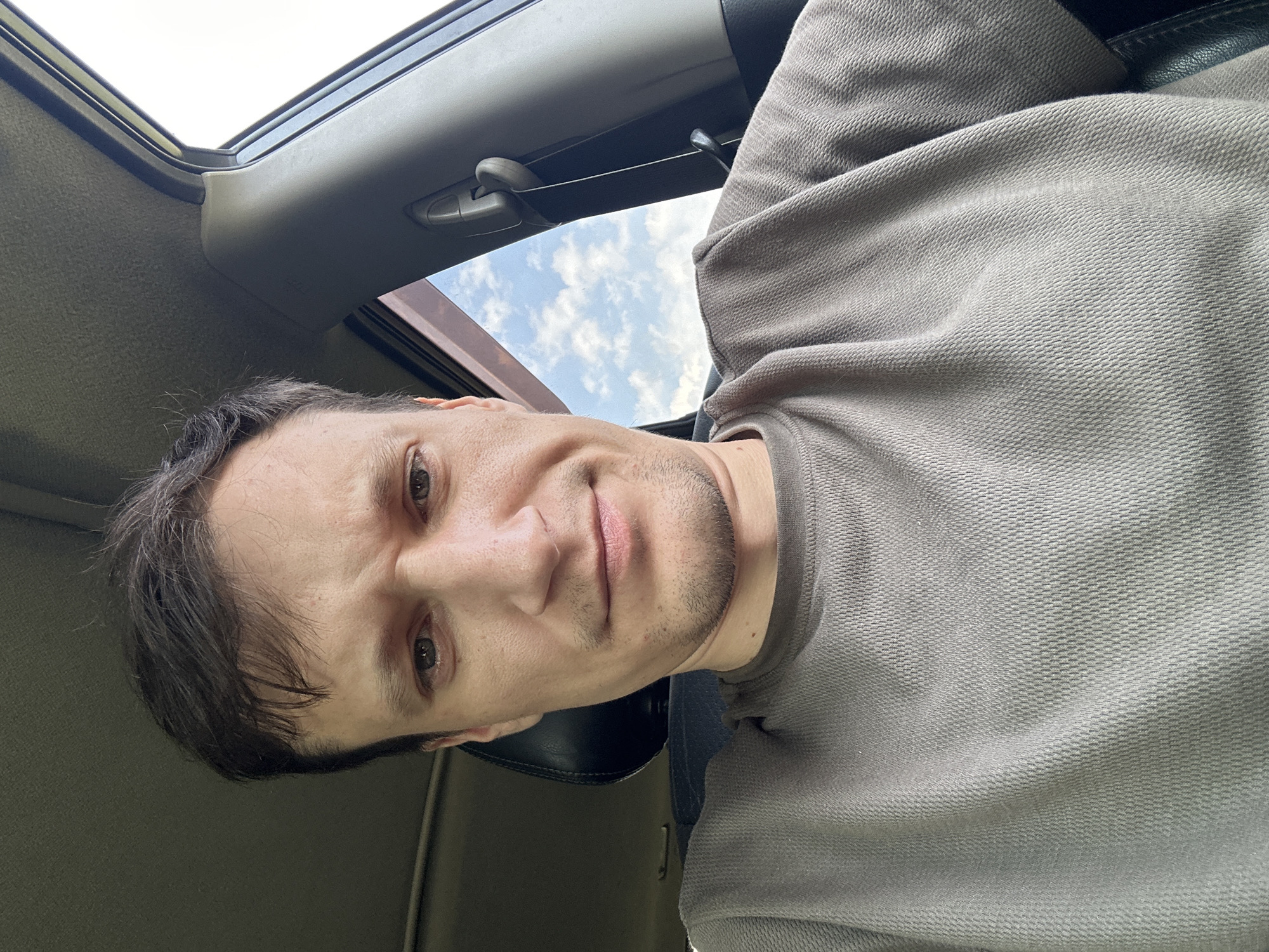 A young Caucasian man with short brown hair and light skin is pictured inside a car, taking a selfie. He is wearing a grey, textured t-shirt. His face is the primary focus, with a slightly furrowed brow and a neutral expression. The background shows the interior of the car, including a headrest and the car's ceiling, as well as a view of the sky with scattered clouds through the window. The overall lighting suggests it is daytime. The image does not contain information regarding the specific location in Makiv, Ukraine.