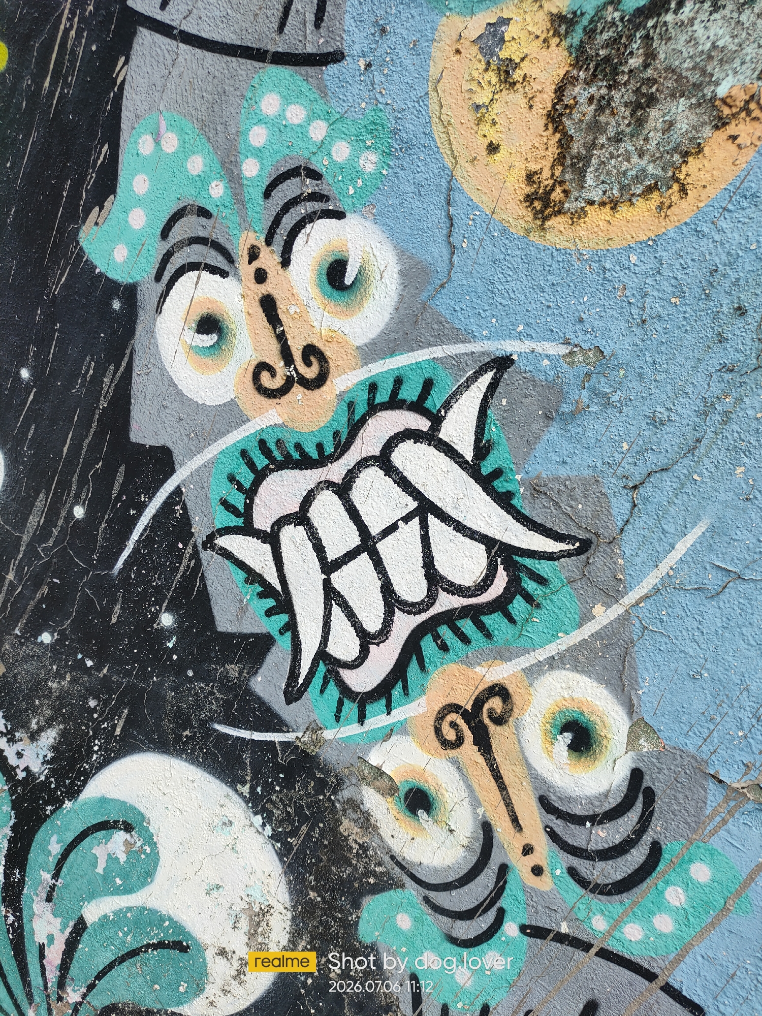 The image shows a close-up of graffiti on a textured, blue wall in Phan Thiết, Vietnam. The graffiti features stylized cartoonish faces with large eyes, expressive eyebrows, and decorative elements. In the upper left quadrant, a face with teal and white polka-dot ear-like structures is visible, with wide white eyes outlined in black and a tan nose with black curlicue details. Below this, a stylized, toothy mouth is depicted within a teal, spiky outline. In the lower right, another face with similar large, round eyes and tan facial features is depicted, with black lines suggesting eyelashes and curved lines below the eyes. The wall itself shows signs of age and wear, with cracks and peeling paint, particularly in the upper right where a textured, yellowish-orange circular shape with dark, rough patches is prominent. White and black paint splatters and streaks are also present, adding to the distressed appearance of the surface. A watermark in the bottom right corner indicates the image was taken by "dog_lover" on "2026.07.06 11:12".