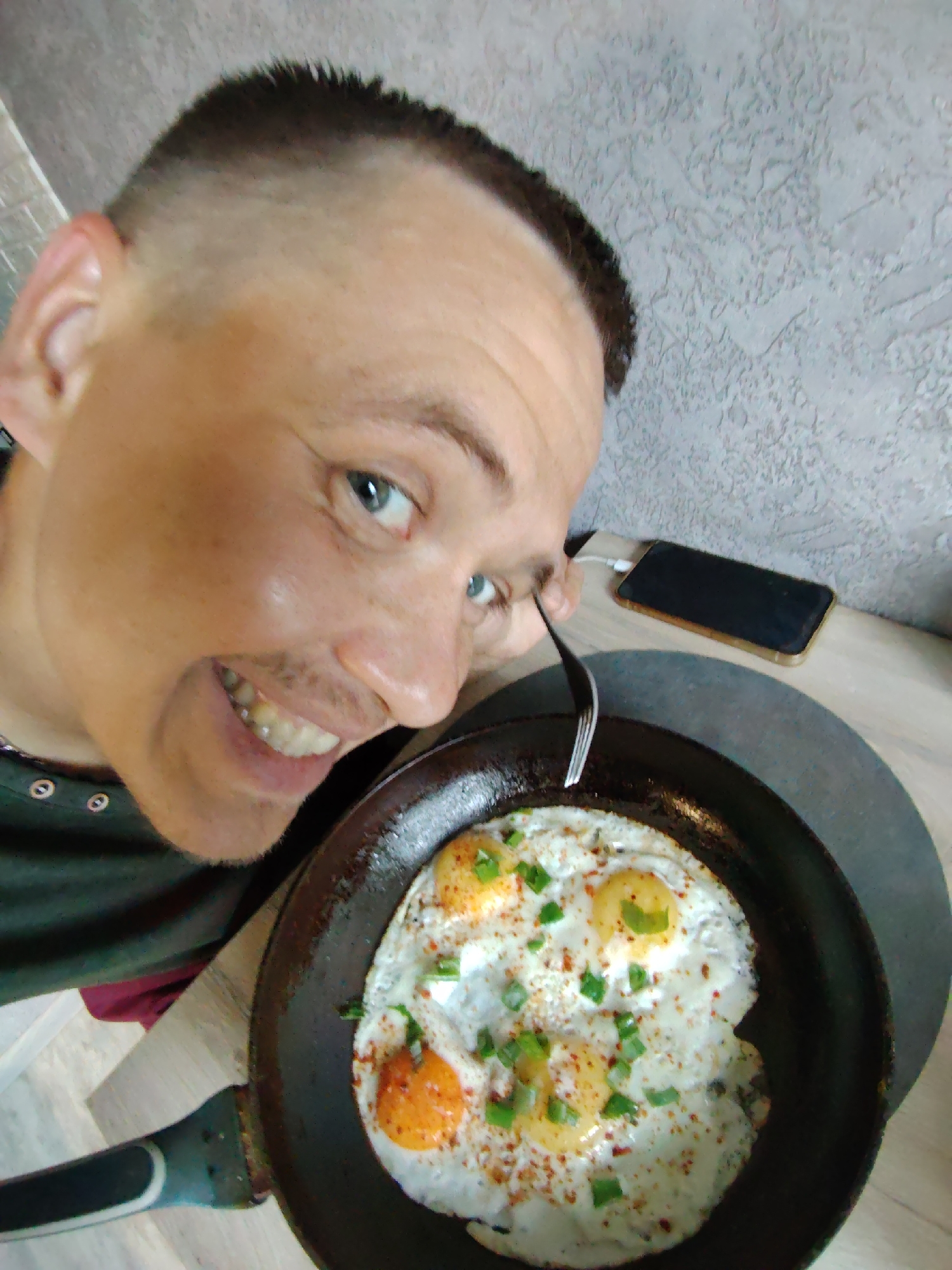 A high-angle medium shot captures a man with a short haircut and a mustache holding a fork and looking towards the camera while a pan of fried eggs rests on a table. The man's head and shoulders fill the left side of the frame, and he is smiling, revealing his teeth. His eyes are blue and he is looking directly at the viewer. He is wearing a dark green shirt with silver buttons. To the right of the man's head, a smartphone with a golden border is placed on a textured gray surface. In the foreground, a black frying pan contains three fried eggs, garnished with chopped green onions and red pepper flakes. The pan is positioned on a dark gray circular placemat, which rests on a light brown wooden table. The background consists of a textured gray wall. The scene appears to be indoors, likely a kitchen or dining area, within a residential setting in Chaikovsky, Russia.