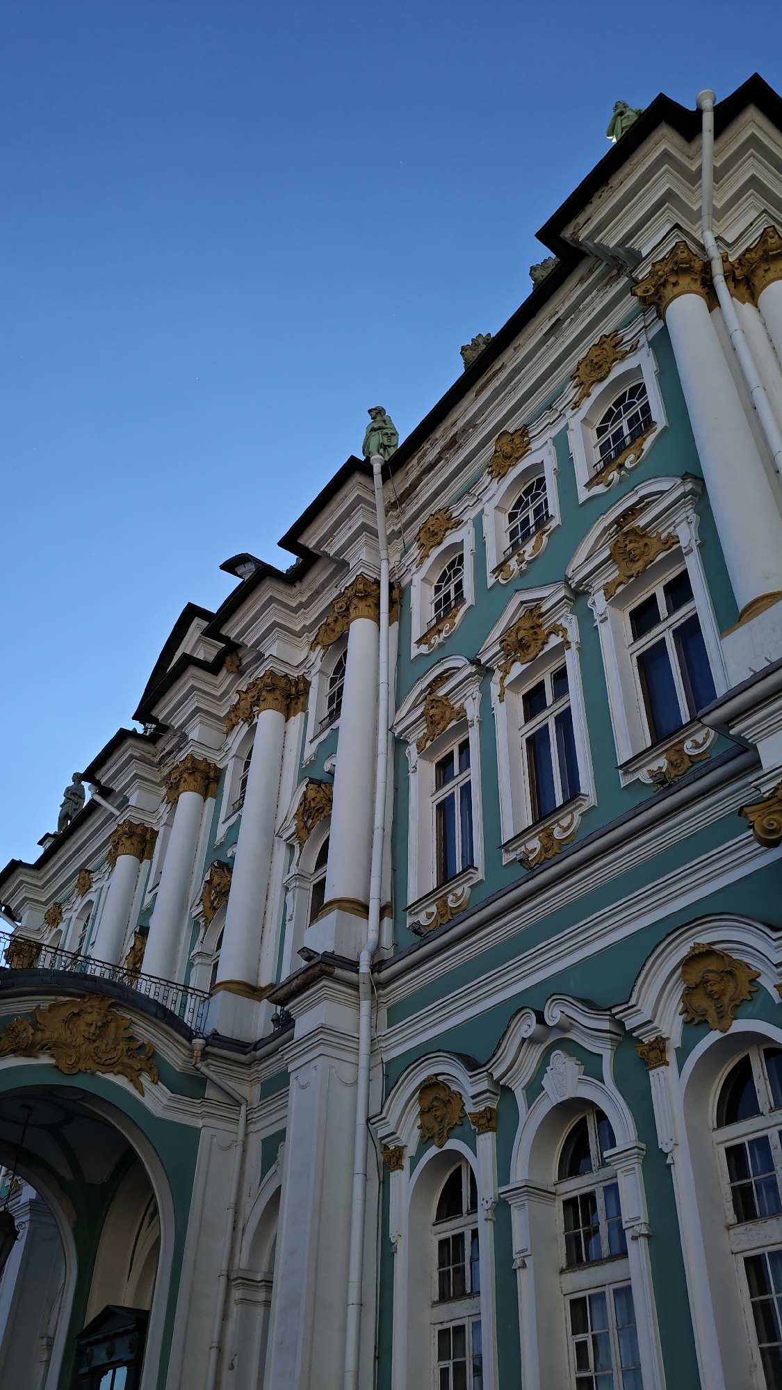 The image shows a low-angle view of the turquoise and white facade of the Winter Palace in Saint Petersburg, Russia, under a clear blue sky. The palace features grand white columns with gold accents, numerous windows with ornate gold surrounds, and decorative sculptural elements. On the roofline, small green statues are visible against the sky. The architectural style is characteristic of the Baroque period, showcasing intricate detailing and a sense of opulence. The building appears well-maintained, and the lighting suggests it is either early morning or late afternoon. No people or specific actions are depicted in the frame. The environment is urban, focusing solely on the exterior of the historic palace.