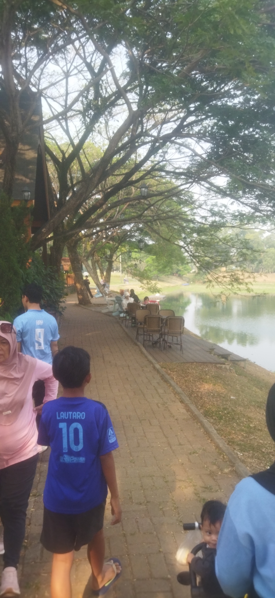 The image depicts a scenic outdoor scene in Depok, Indonesia, likely a park or recreational area adjacent to a body of water. In the foreground, several individuals are walking along a brick pathway. A young boy wearing a blue t-shirt with "LAUTARO 10" on the back and black shorts is walking towards the camera. To his left, a woman in a pink headscarf and pink long-sleeved shirt is also walking. Further back, a man in a light blue t-shirt with "HAY 9" on the back is visible.
The pathway leads towards a water body, possibly a lake or pond, on the right side of the image. Along the edge of the water, there is a wooden deck with several empty chairs and tables set up, suggesting an outdoor dining or seating area. In the distance, more people are seated at the tables, and the background features lush green trees and vegetation, characteristic of a park environment. The overall atmosphere appears relaxed and leisurely, with the natural beauty of the surroundings being a prominent feature.