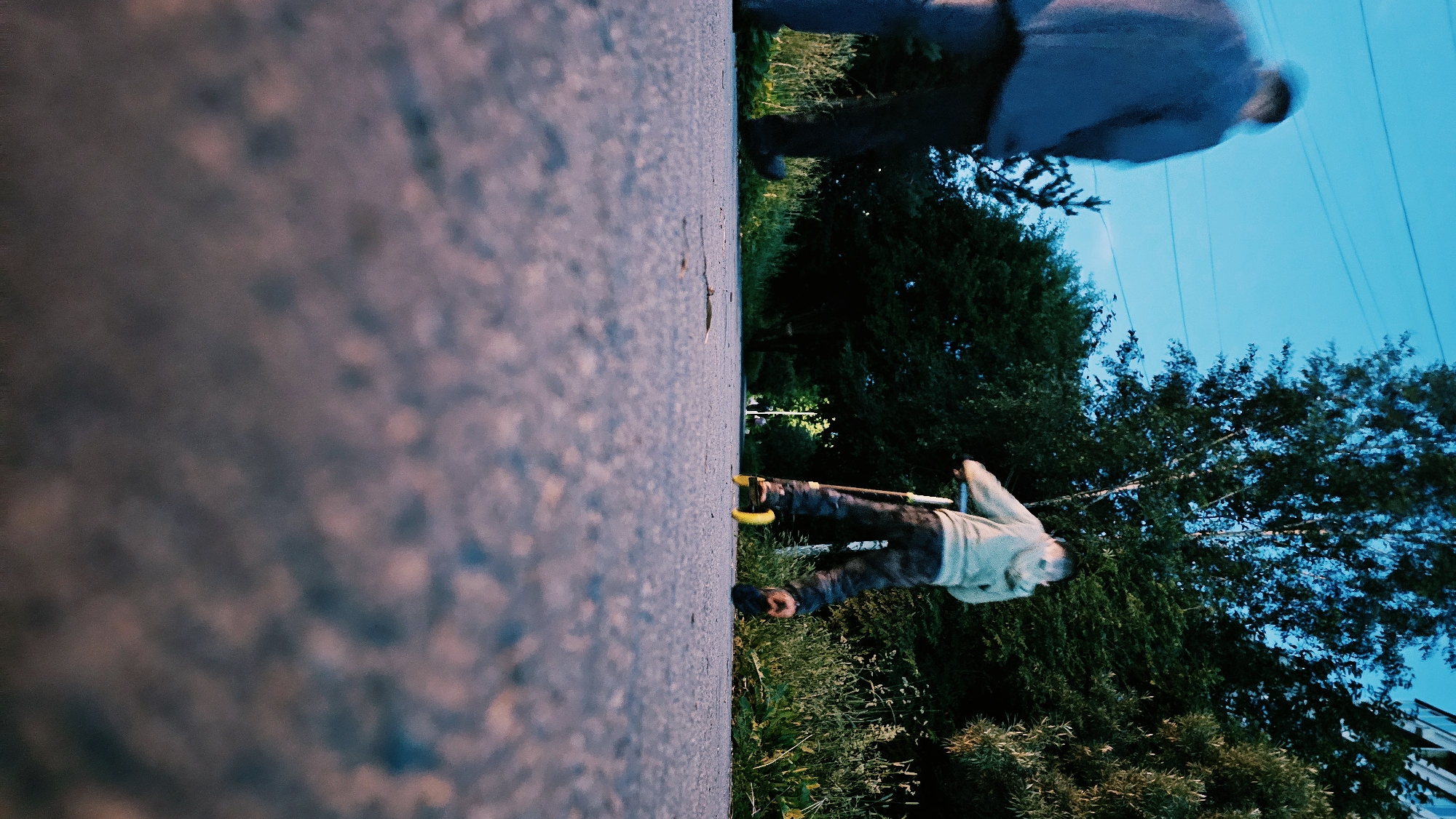 The image depicts a person on a scooter, seen from a low-angle perspective. The scooter has bright yellow accents on its wheels. The person is wearing dark pants and a light-colored hooded jacket. The scene is outdoors, with a paved surface in the foreground and lush green trees and foliage in the background. The sky is a clear, vibrant blue, suggesting it is daytime, possibly late afternoon or early evening given the lighting. The image is oriented vertically, with the ground running along the left side and the sky and trees occupying the right side. The focus is sharp on the scooter and the person's legs, with the foreground and background slightly blurred. There are no visible text elements in the image. The overall mood is active and casual, suggesting a moment of outdoor recreation.