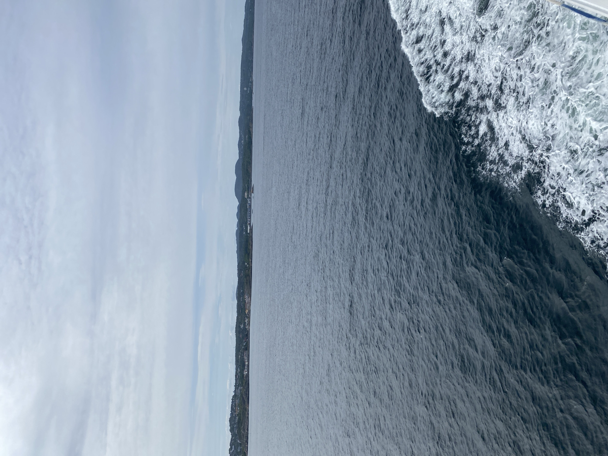 This is a photograph taken from a boat at sea, looking towards a coastline. The perspective is tilted, giving a sense of movement and depth. The foreground on the right is dominated by the churning white wake of the boat, indicative of a vessel in motion. The wake transitions into the dark, choppy waters of the sea.
In the mid-ground, a distant coastline is visible. It is characterized by rolling, tree-covered hills that slope down to the water. Scattered along the coastline are hints of human settlement, with buildings and structures clustered in some areas. The overall impression of the coastline is one of natural beauty with human presence.
The background is a hazy sky with soft, white clouds. The lighting suggests it could be daytime, possibly late morning or afternoon, with overcast or partly cloudy conditions. The sky is a pale blue, blending with the lighter tones of the clouds.
The scene evokes a feeling of travel and exploration. The motion of the water and the distant view of land create a sense of journeying. The location cues, such as the hilly, forested coastline, are consistent with a Norwegian setting like Stavern, known for its scenic coastal landscapes. There is no visible text in the image.