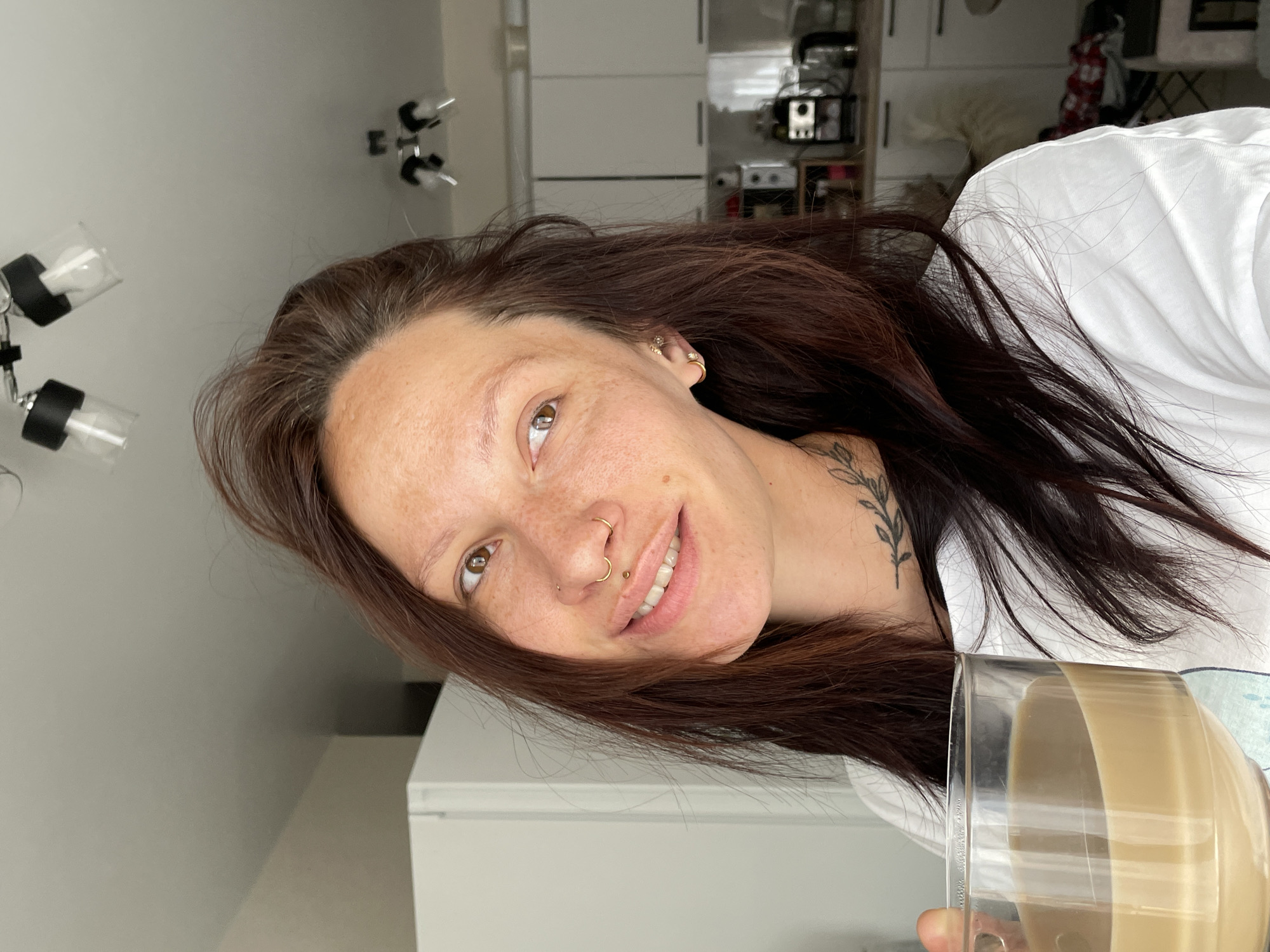 A fair-skinned woman with reddish-brown hair, freckles, and a nose piercing smiles at the camera while holding a glass of a light-colored beverage. She wears a white shirt and has a leafy tattoo on her neck. The background shows a kitchen interior with white cabinets and a counter where kitchen appliances are situated. A lighting fixture is mounted on the ceiling. The scene is indoors and well-lit. There is no indication of the location being in Pervomaysky, Russia.