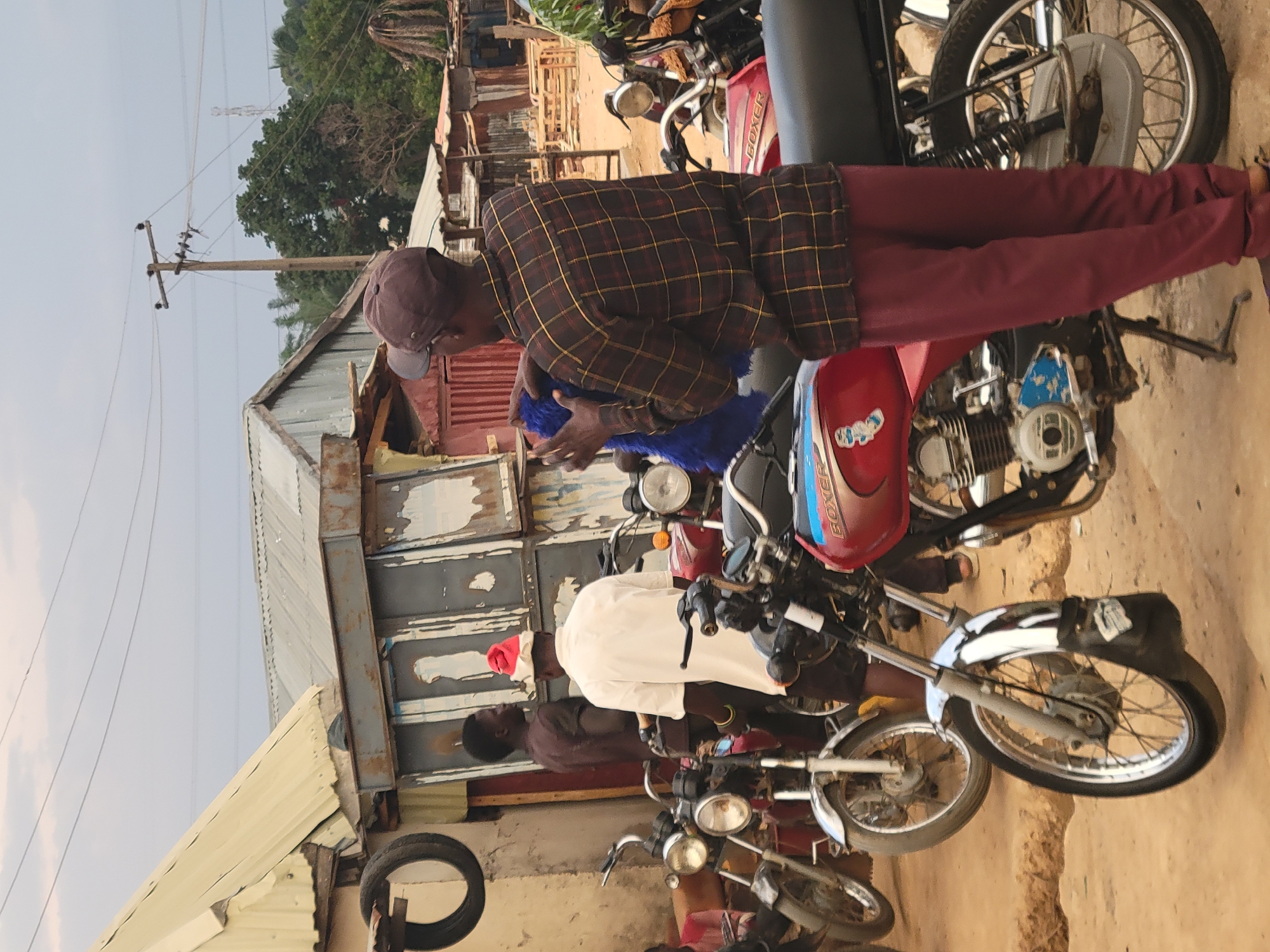 The image depicts a street scene in Abuja, Nigeria, with a group of men gathered around several motorcycles. One man, wearing a plaid shirt and maroon pants, is standing near a red motorcycle with a blue seat cover. Another man in a white shirt and a red cap is standing near the motorcycles. The background shows a building with corrugated metal walls and roofing, along with power lines and trees.