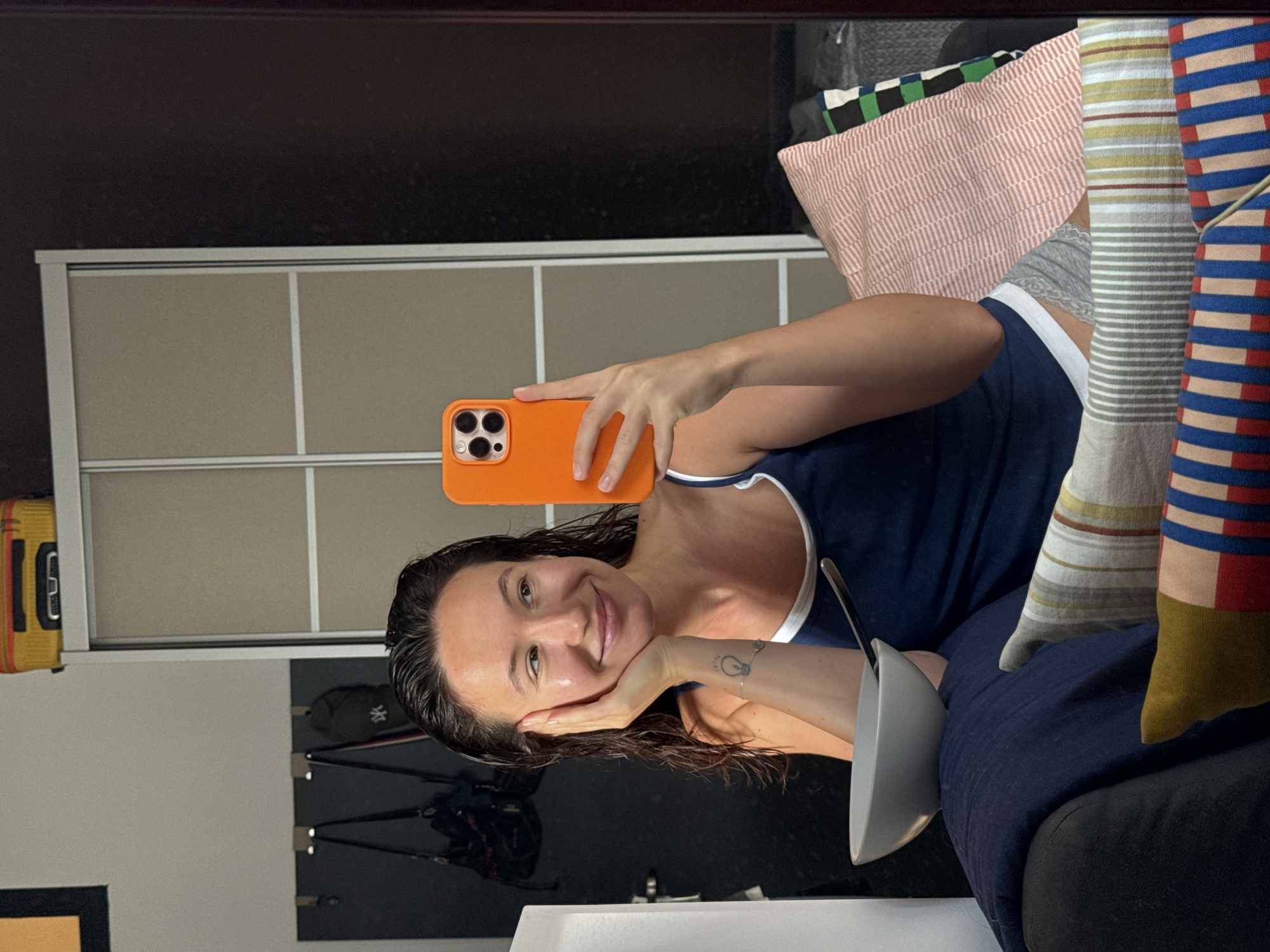 A young woman with wet, dark hair is taking a selfie in a mirror, resting her left hand on her cheek and holding her orange smartphone in her right hand. She is wearing a navy blue tank top with white trim and grey shorts. A light grey bowl with a spoon is visible in the foreground on her lap, which is covered by a dark blue blanket. Behind her, there are several pillows with striped patterns and a closet with sliding doors. The scene appears to be indoors, possibly in an apartment. The image does not contain any identifiable landmarks or features that would suggest a specific location within Tel Aviv, Israel.