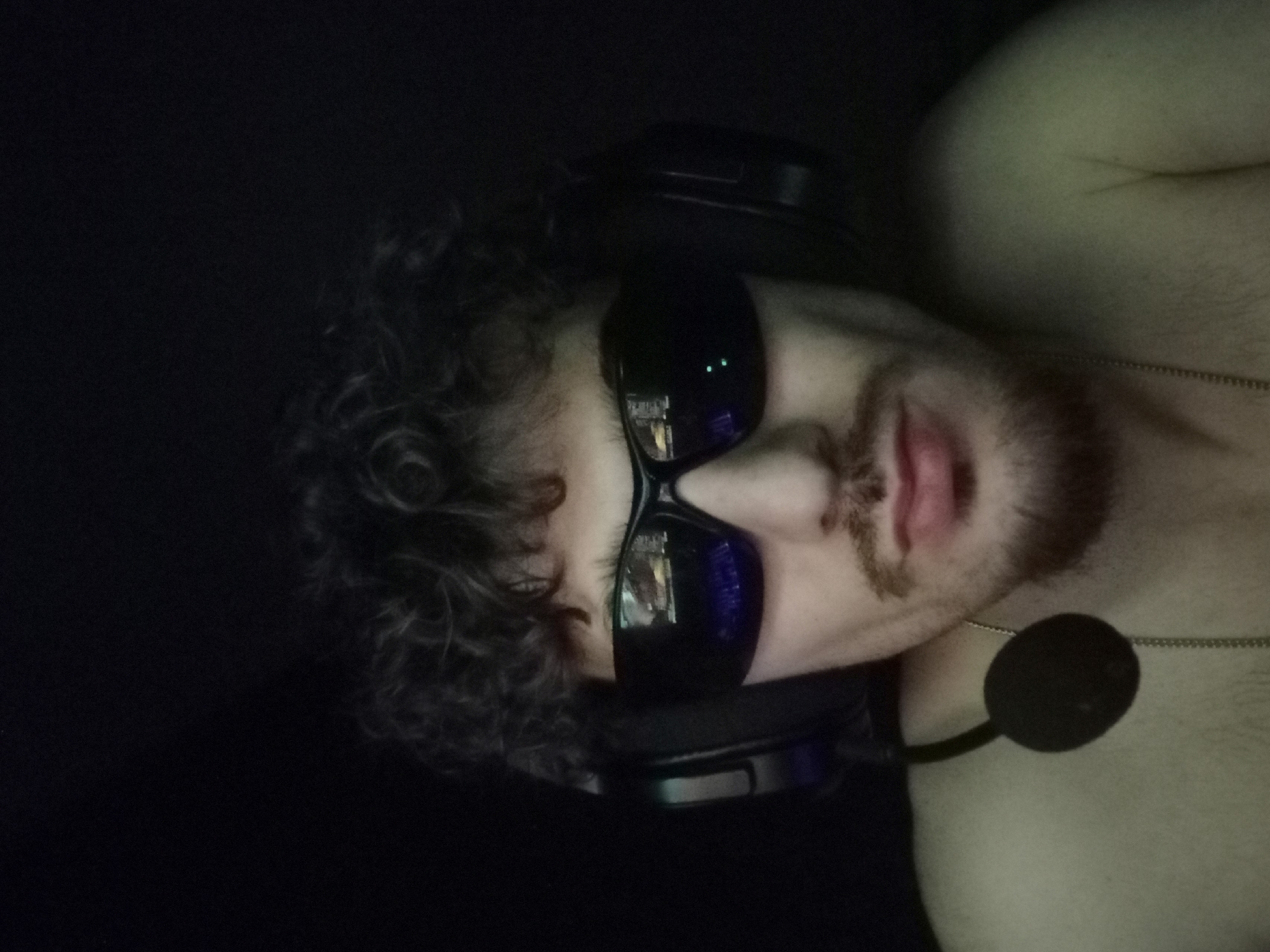 The image is a close-up, head-and-shoulders portrait of a young man wearing black sunglasses and headphones. The man has curly dark hair and a mustache and goatee. He is shirtless. The background is dark, with some subtle green lighting on the right side of the image. The sunglasses reflect what appears to be a computer screen, showing some colorful graphics. A microphone is attached to the headphones and hangs near his neck. A thin silver chain is visible around his neck. There are no discernible elements in the image that would indicate a specific location within Parvomay, Bulgaria, or any city.
