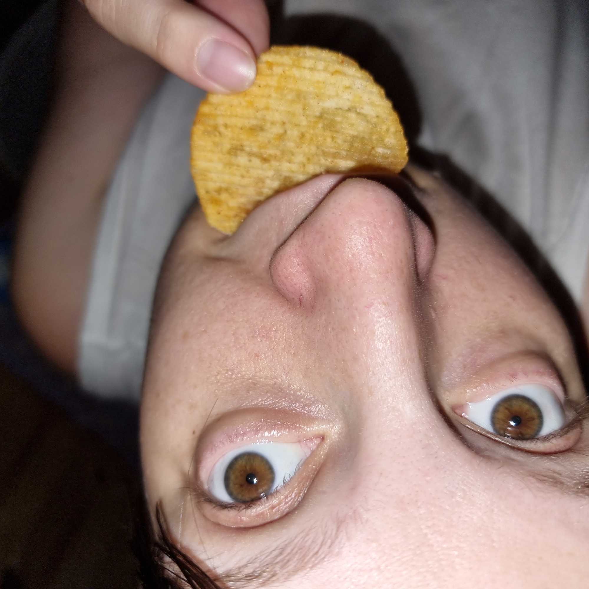 In the city of Dunnae-myeon, South Korea, a close-up, eye-level shot captures a person's face as they eat a potato chip. The individual, with light skin and brown eyes, is shown from just below the forehead to the chin. Their left eye is slightly more visible than the right, which is partially obscured by the chip. The potato chip, ridged and golden brown, is held between the person's thumb and forefinger, and is positioned near their mouth. The background is dark and out of focus, suggesting an indoor setting. The person is wearing a light-colored garment, possibly a tank top or t-shirt. The lighting is direct, highlighting the texture of the skin and the details of the chip.