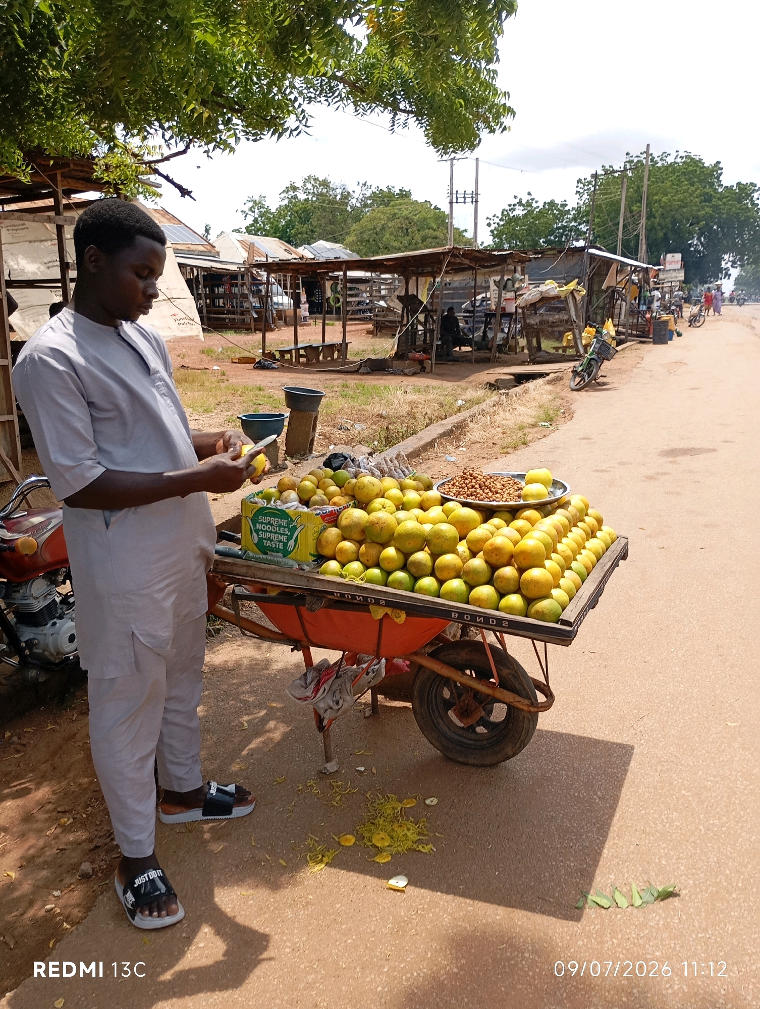 A young Black male, dressed in a light gray traditional outfit and black and white slide sandals, stands beside a cart laden with oranges and nuts in a market setting in Shendam, Nigeria. He is in the process of peeling an orange with a small knife. The cart, which appears to be a repurposed wheelbarrow, is filled with a large display of oranges, some in a yellow box labeled "SUPREME NOODLES, SUPREME TASTE," and a metal bowl containing nuts. Behind the vendor and his cart, market stalls with corrugated metal roofs are visible, constructed from wood and fabric. A motorcycle with an orange fuel tank is parked to the left of the cart. In the background, other market activity is suggested with indistinct figures and structures. The ground is unpaved, with the cart resting on a sandy, dirt surface. The scene is outdoors, with overhead trees providing partial shade. The sky above is partly cloudy.