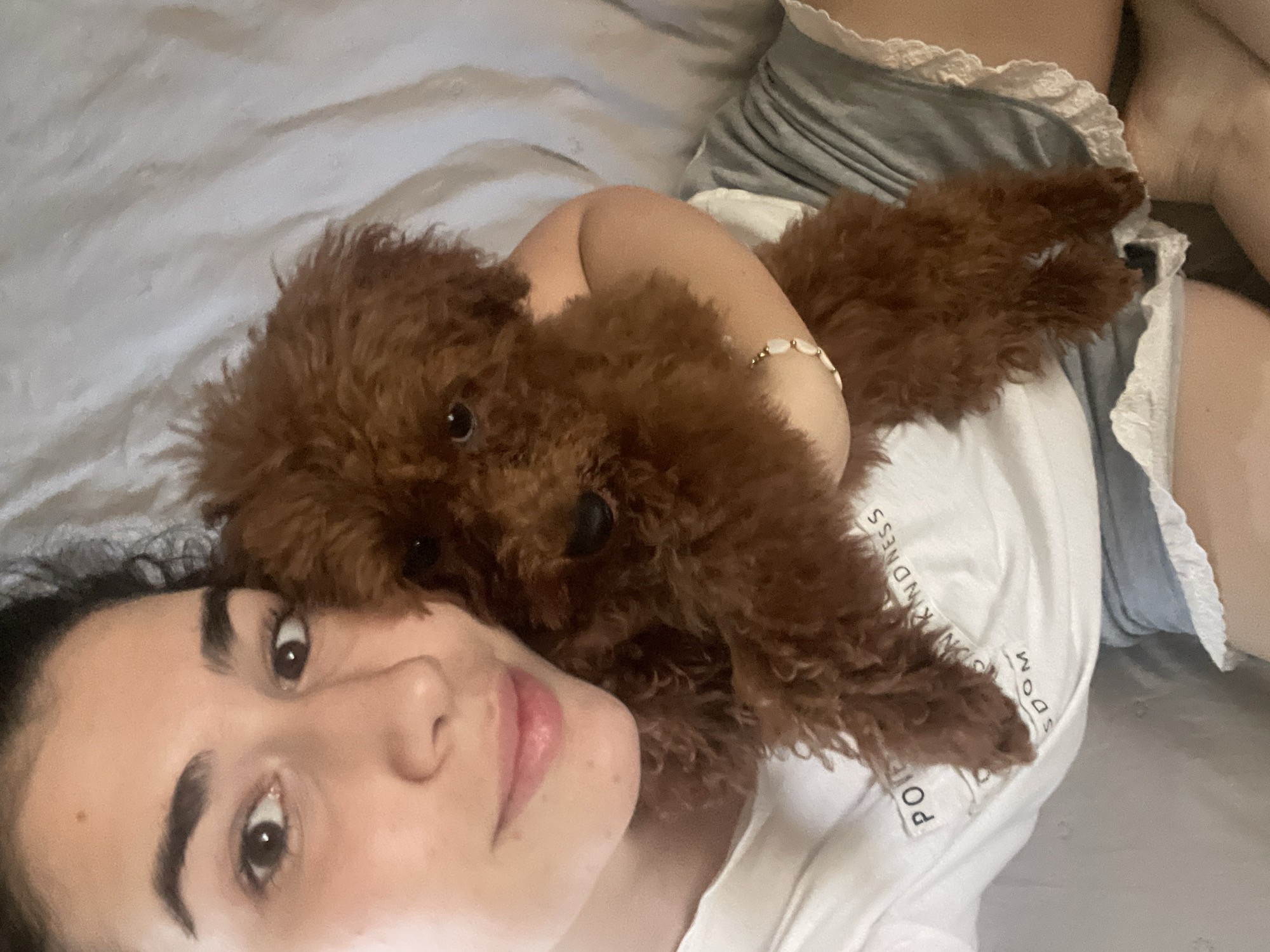 A selfie taken from an eye-level perspective features a woman lying down with a small, brown poodle resting on her chest. The woman has dark hair and eyebrows, light skin, and is wearing a white t-shirt. The poodle's fur is dense and curly. The background consists of rumpled, light-colored bedding. The location is indiscernible and cannot be placed in Sofia, Bulgaria based on the provided image.
