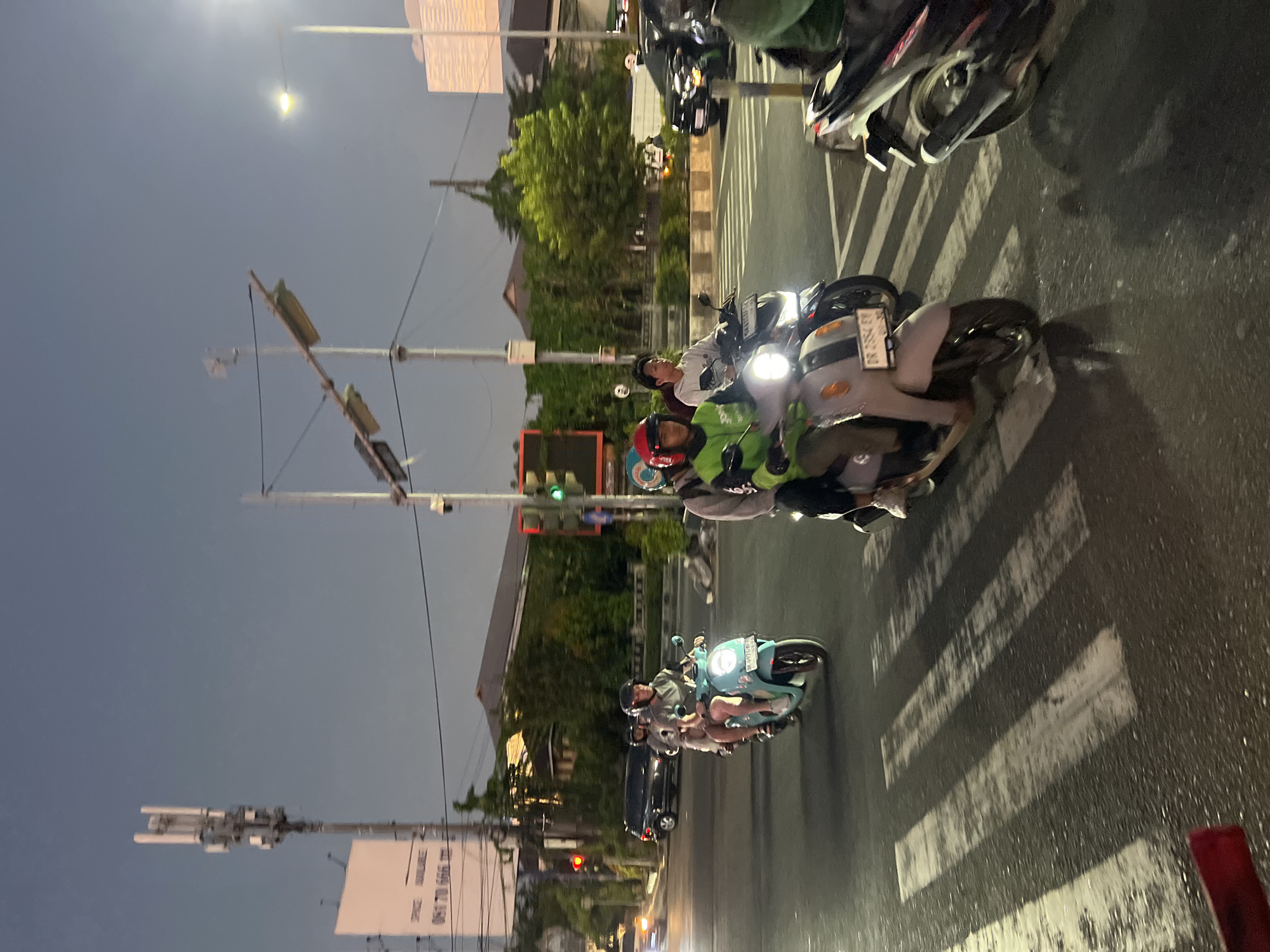 At dusk on a city street in Denpasar Barat, Indonesia, several motorbikes are seen on a wet asphalt road, with some vehicles in motion at a crosswalk. A man and a woman, both wearing helmets, ride a light blue scooter, with the woman in the back wearing a red helmet. Another motorbike, ridden by a man in a green jacket and a red helmet, is positioned ahead of them, with a second person behind him wearing a grey sweatshirt. The man in the green jacket appears to be a delivery driver. In the background, a black car is stopped at a traffic light, with another car visible further down the road. There are streetlights, traffic signals, and a telecommunications tower, indicating an urban environment. The sky is a deep blue, transitioning into evening. The road surface shows reflections of lights and the white stripes of a pedestrian crossing are clearly visible.