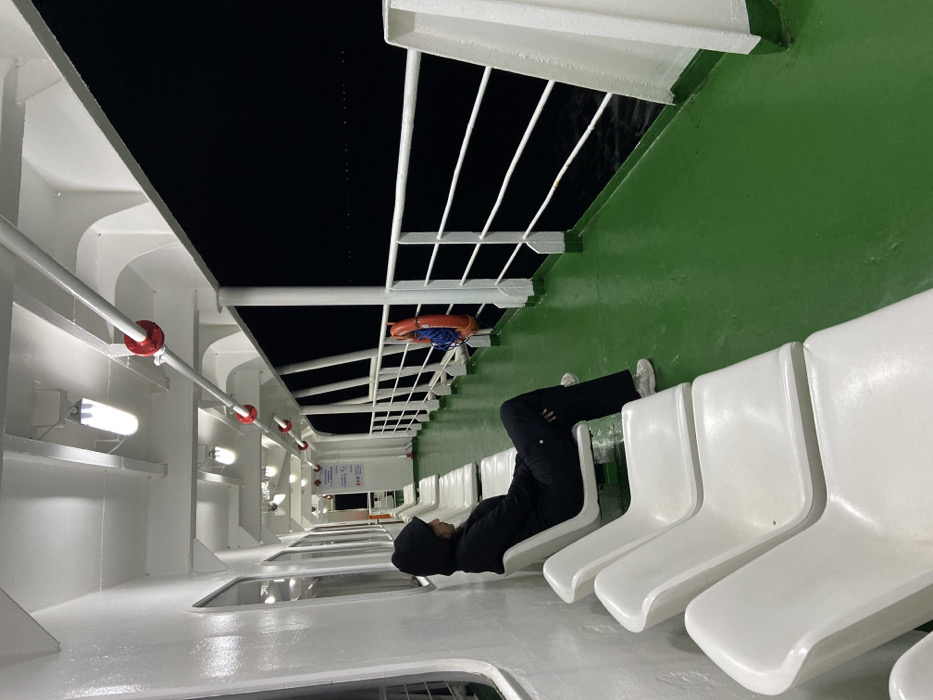 The image depicts a person reclining across four white chairs on what appears to be the deck of a ship or ferry. The person is wearing a dark hooded sweatshirt and pants with white sneakers. Overhead, there is a metal railing, a life preserver, and views of the dark sea. The floor is green. The ship's structure is predominantly white. The perspective suggests the photograph has been rotated.