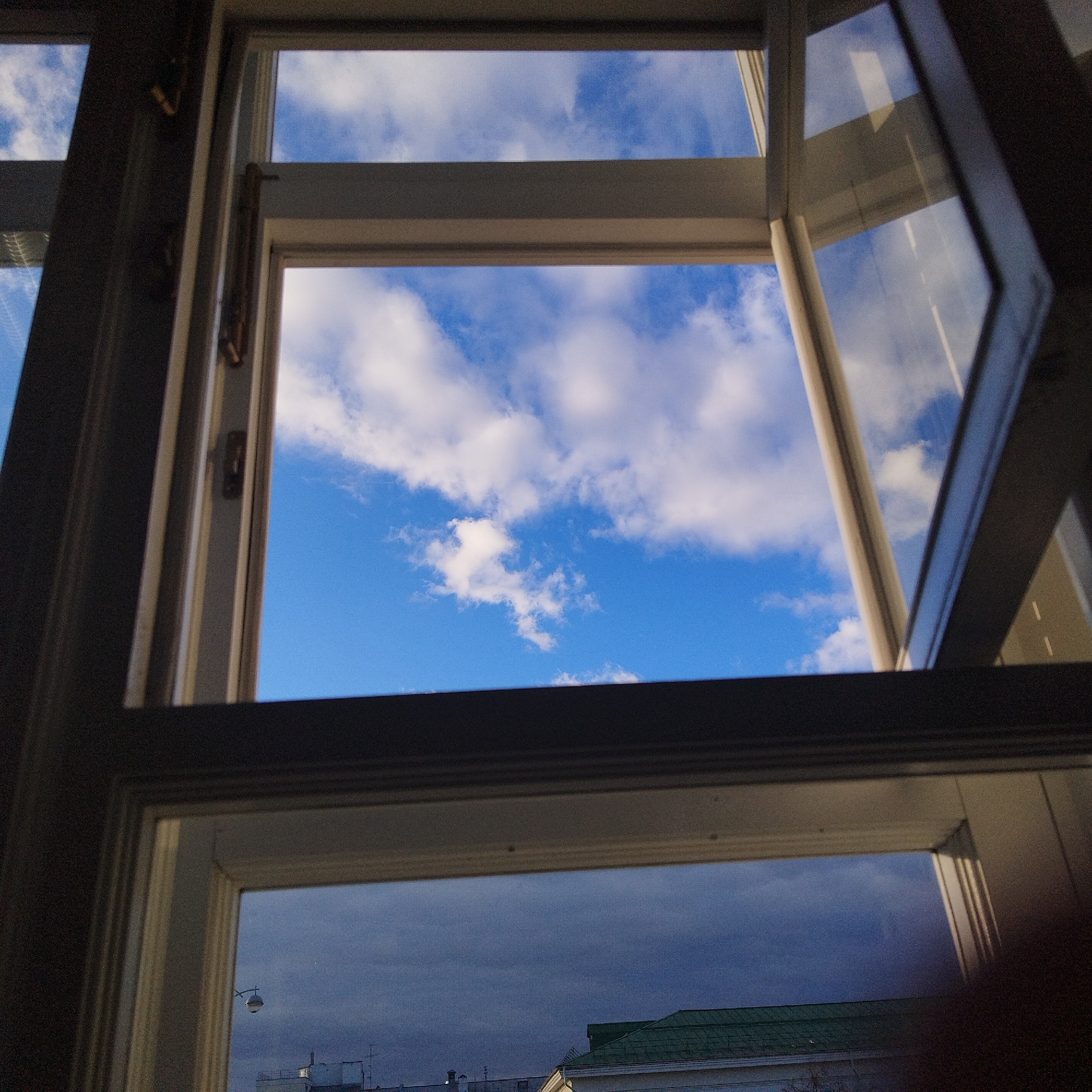 The image shows a view through a partially open, multi-paned window. The top pane reveals a bright blue sky with scattered white clouds. The window is white with visible hardware. The bottom pane displays a darker sky, suggesting a change in weather or time of day. The faint outlines of rooftops and a street light can be seen through the lower pane, indicating an urban setting. Based on the architectural styles present, this could be in Yekaterinburg, Russia. The scene is static, depicting a moment captured through the window.