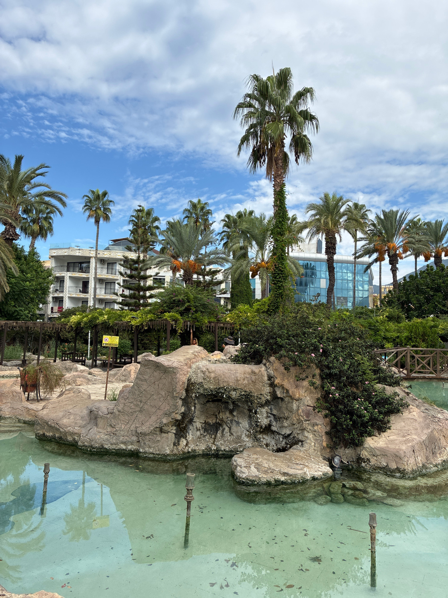 The image depicts a resort area with a swimming pool or water feature in the foreground. The water is clear and turquoise, with a few fallen leaves visible on the bottom. Several metallic poles, possibly part of a fountain system, protrude from the water. Large, artificial rock formations are situated on the edge of the water, covered in moss and some greenery. A small wooden bridge crosses over a portion of the water in the mid-ground to the right.
Behind the water feature and rock formations, a shaded area with wooden pergolas and benches is visible, suggesting a seating or relaxation zone. A yellow warning sign is affixed to one of the pergola posts. A small, brown, animal-like statue or ornament is present on the rocks near the pergola.
The background features lush greenery, including numerous palm trees of various sizes. Behind the trees, a multi-story white hotel building with balconies is visible, followed by a modern building with a glass facade that reflects the sky and surrounding trees. The sky is blue with scattered white clouds. The overall setting appears to be a resort or hotel complex in Alanya, Turkey, designed to resemble a tropical oasis with its abundant palm trees and water features. The image captures a daytime scene under a bright, cloudy sky.