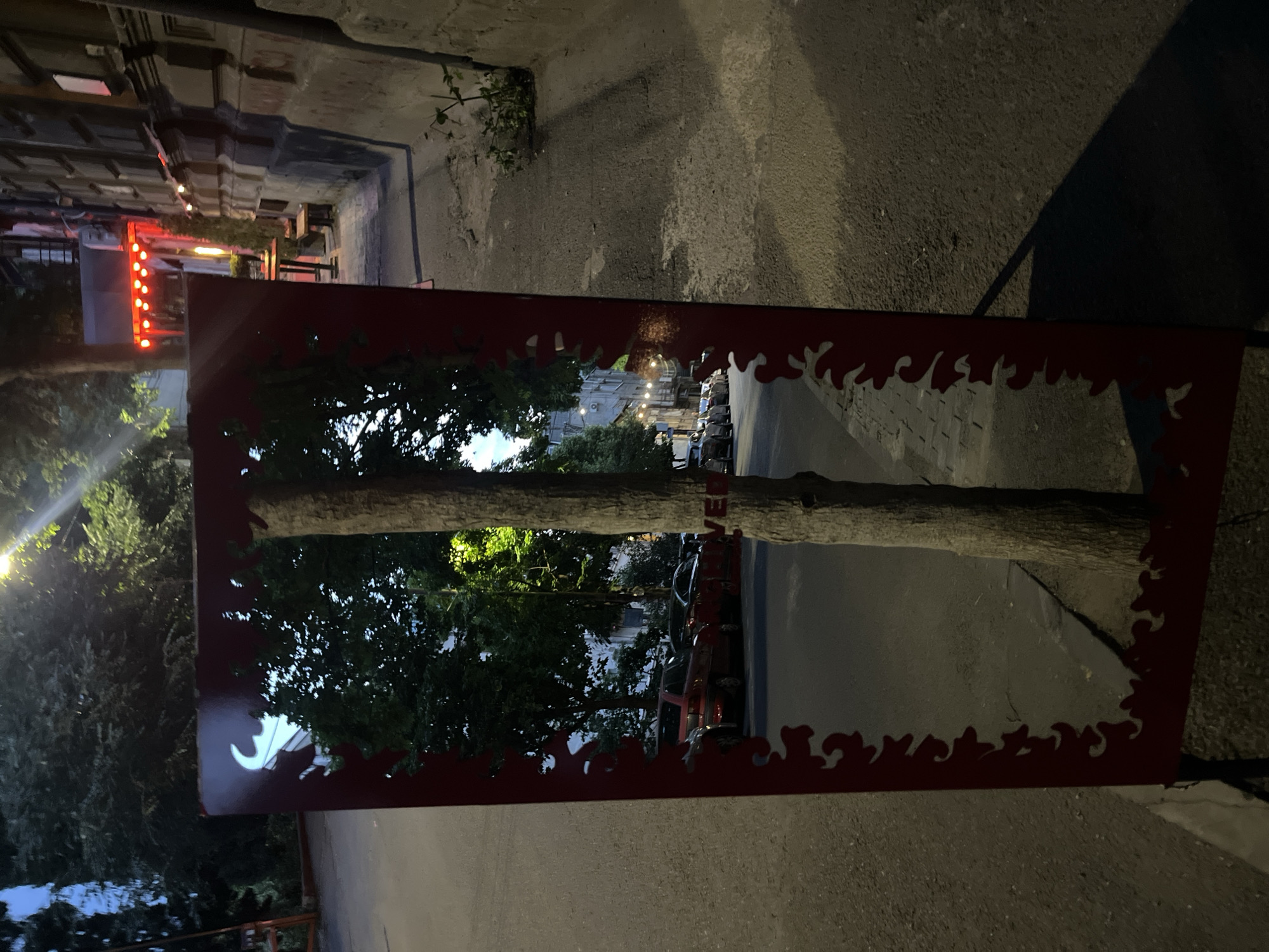 A mirror with a flame-shaped red frame reflects a tree trunk, branches, and street scene in Tbilisi, Georgia. The mirror is placed on the sidewalk. The tree trunk passes directly through the center of the mirror. In the reflection, cars are parked along the street, and lights are visible in the distance. The sidewalk is concrete. The frame has a jagged, flame-like pattern cut out. The image is rotated. Some architectural detail and string lights are visible in the background to the left.