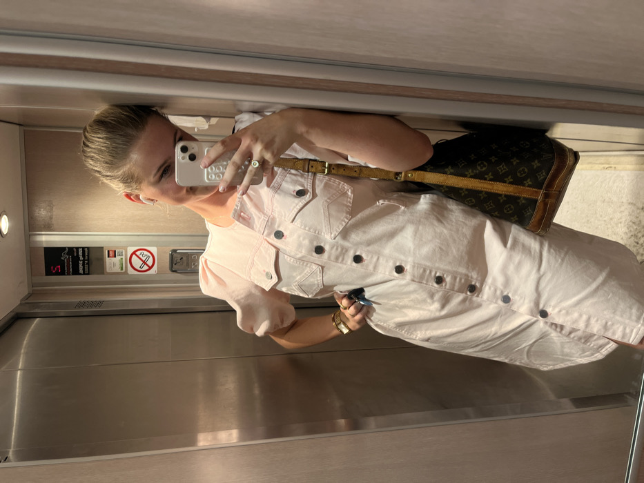 A woman in a pink button-down dress takes a selfie in an elevator. She holds a smartphone with a white case. A brown Louis Vuitton bag is slung over her shoulder. She wears a gold watch on her wrist and a ring on her finger. The elevator's interior features stainless steel doors, signs, and control panels. The lighting is bright and even.