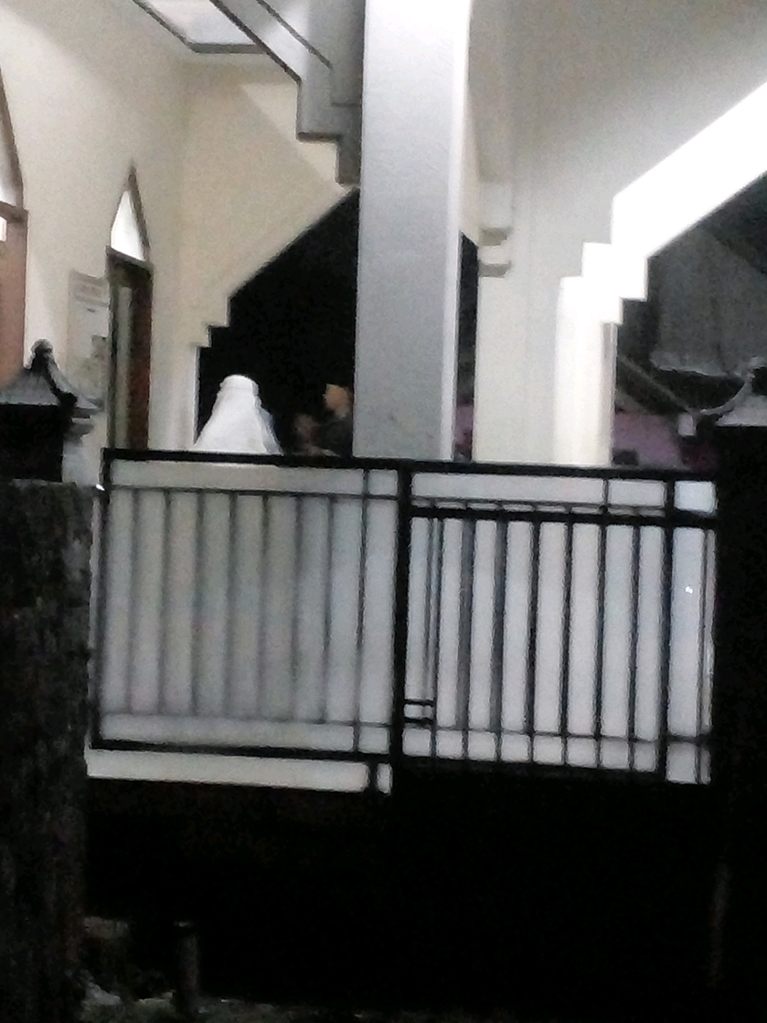 The image depicts a dark, out-of-focus scene at night in Pungangan, Indonesia. In the foreground, a black metal gate with vertical bars and a horizontal railing obstructs the view. Beyond the gate, a person wearing a white hijab stands partially visible. The background suggests an interior space with a staircase and walls. The lighting is dim, and the overall quality of the image is poor, making detailed observation difficult.