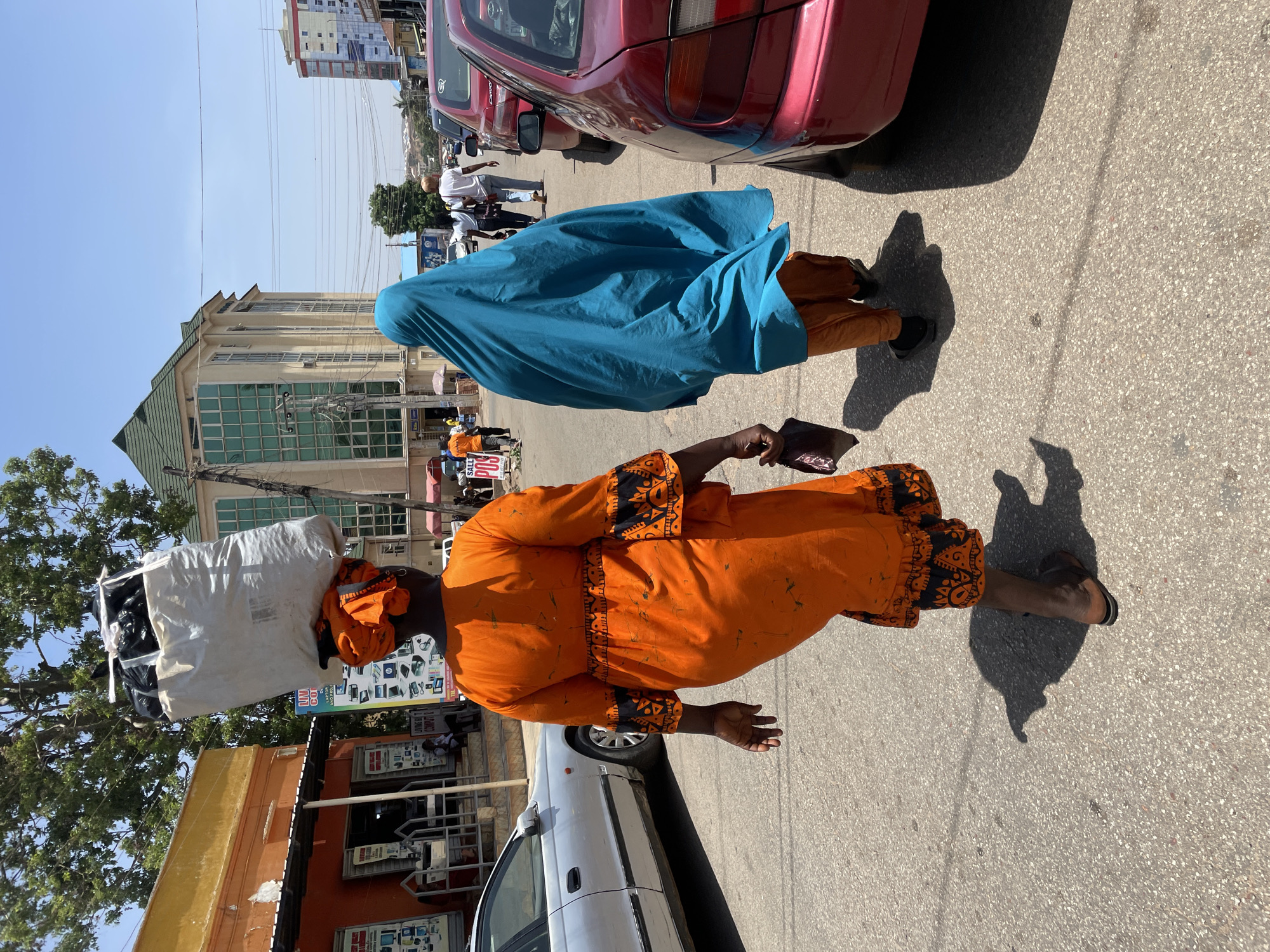 The photograph captures a street scene in Jos, Nigeria. In the foreground, two women are walking along the asphalt road. The first woman is wearing a vibrant orange dress with black geometric patterns and carrying a large white sack on her head. The second woman is clad in a blue garment that covers her head and body.
A red car is partially visible at the top of the image. Buildings with multiple stories and windows are situated in the background. A silver car is parked along the street. Other people are walking in the distance, and commercial signage is visible on the buildings. The scene is bathed in bright daylight.