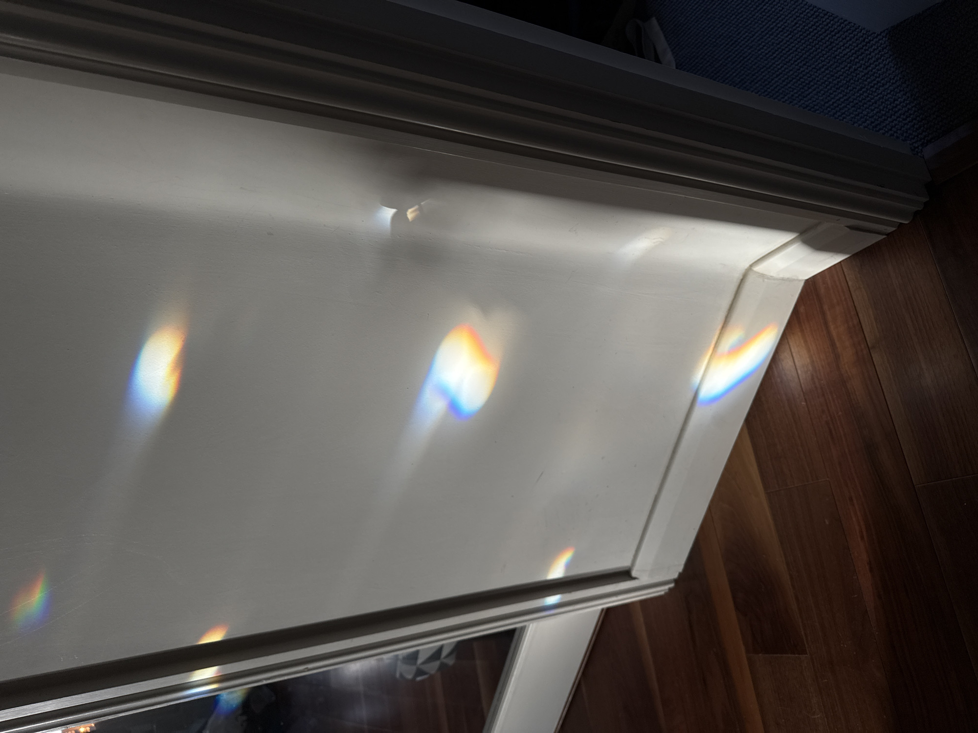 A close-up indoor shot shows a section of what appears to be a white cabinet or mantelpiece. The surface is illuminated by bright, rainbow-like light refractions, creating circular shapes with vivid spectral colors. The top of the cabinet or mantelpiece has a decorative trim with multiple levels. Parts of the background are visible: a dark gray or blue carpeted area at the top and wood flooring on the right side. The overall lighting suggests a bright light source causing the refractions. The location in the city of East Suffolk, United Kingdom, cannot be determined from the image.