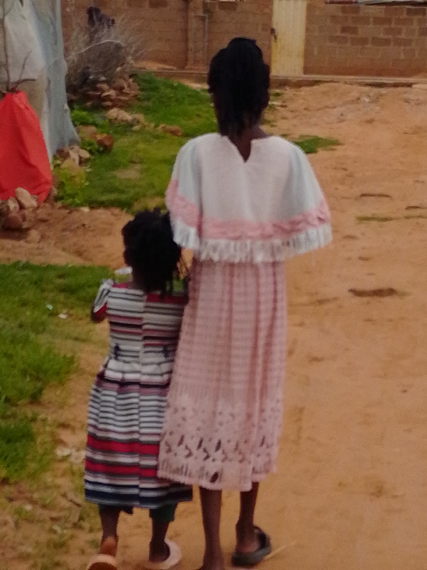 Two young Black females, viewed from behind, stand in an outdoor, dusty environment with some patchy green grass and a brick building in the background. The taller female, on the right, wears a pink and white patterned dress with a white ruffled trim around the neckline and sleeves. The shorter female, on the left, wears a horizontal striped dress in various shades of blue, red, and white. The ground is a light brown, sandy dirt. There is a red tarp or cloth draped over some objects on the left side of the image. The building in the background is constructed of reddish-brown bricks and has a yellow door. The lighting suggests it is daytime.