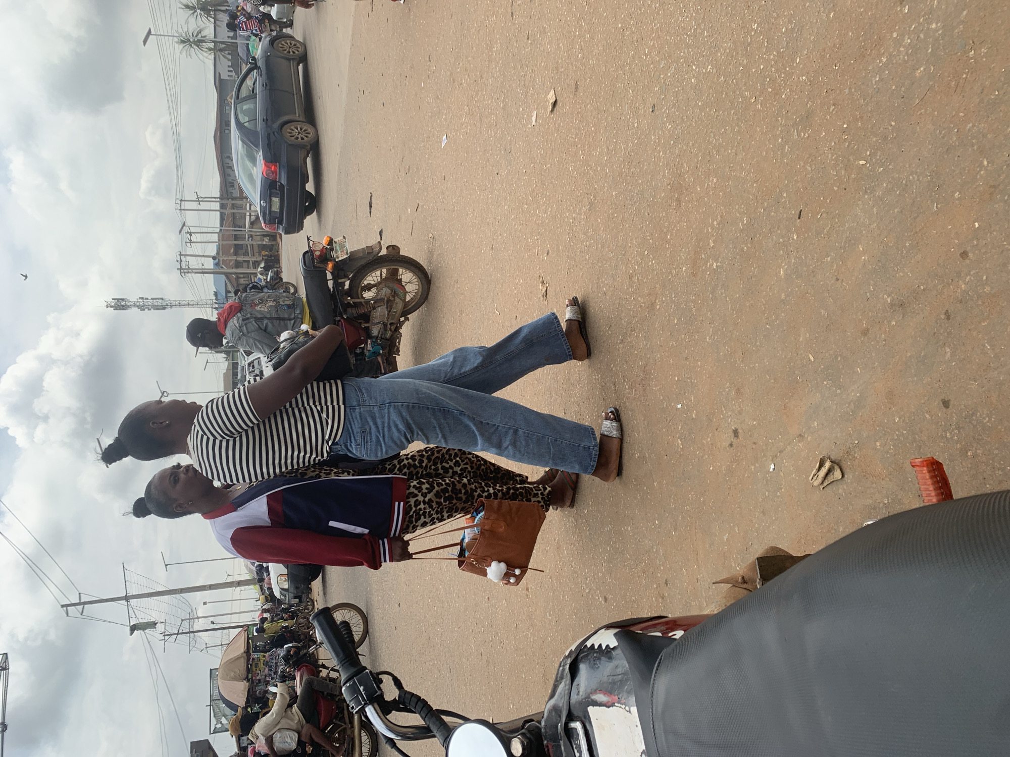 Two women stand on a dusty road in what appears to be an urban environment in Shagamu, Nigeria. The woman in the foreground wears a white and black striped shirt, blue jeans, and sandals. She is holding a brown purse with a white fluffy keychain. The woman behind her wears a red, white, and blue jacket over a leopard print outfit. She is also wearing brown sandals. They are standing near a parked motorcycle.
In the background, a dark-colored sedan is parked on the side of the road. Further back, several other motorcycles and a person in a red jacket are visible, suggesting a busy street or market area. Power lines and utility poles are prominent in the scene, indicating an developed area. The sky is cloudy, suggesting it could be overcast or a transition in weather. The overall impression is a snapshot of everyday life on a street in Nigeria.