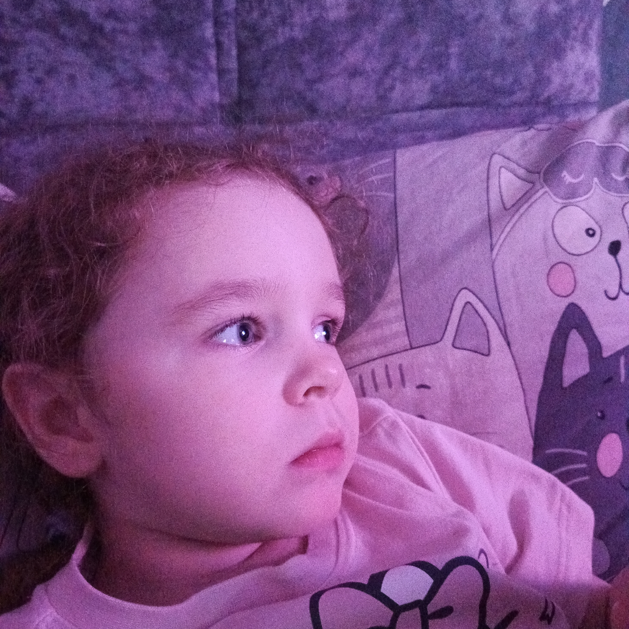 A young child is lying on a pillow and blanket. The blanket is patterned with cat faces. The child is wearing a pink shirt and is looking to the left of the frame. The location is not discernible from the image.