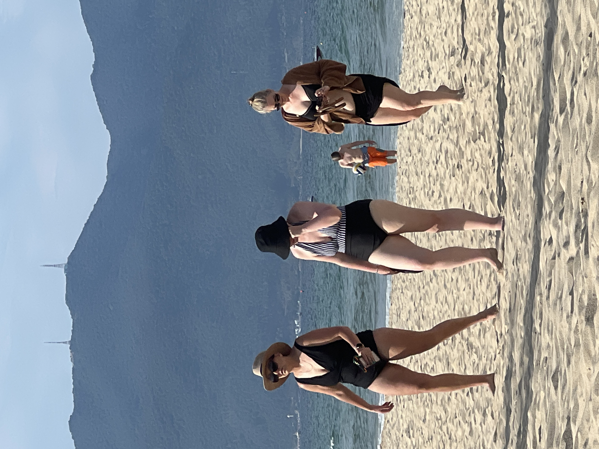 The image captures a scene on a sandy beach in Đà Nẵng, Vietnam, with the Sơn Trà Peninsula mountains forming a backdrop. Three women in swimwear are walking towards the right of the frame on the sand. The woman on the left wears a black one-piece swimsuit and a straw hat with sunglasses, holding an object in her hand. The woman in the middle is in a black and white striped one-piece swimsuit and a black bucket hat. The woman on the right is in a black bikini bottom and a black bikini top, with a brown cardigan and sunglasses, and is looking at her phone. In the background, a man in orange shorts is visible near the water, holding a volleyball. The sea stretches to the horizon, and the Sơn Trà Peninsula mountains are visible in the distance, with two distinct towers on its peak. The sky is a clear, pale blue.
