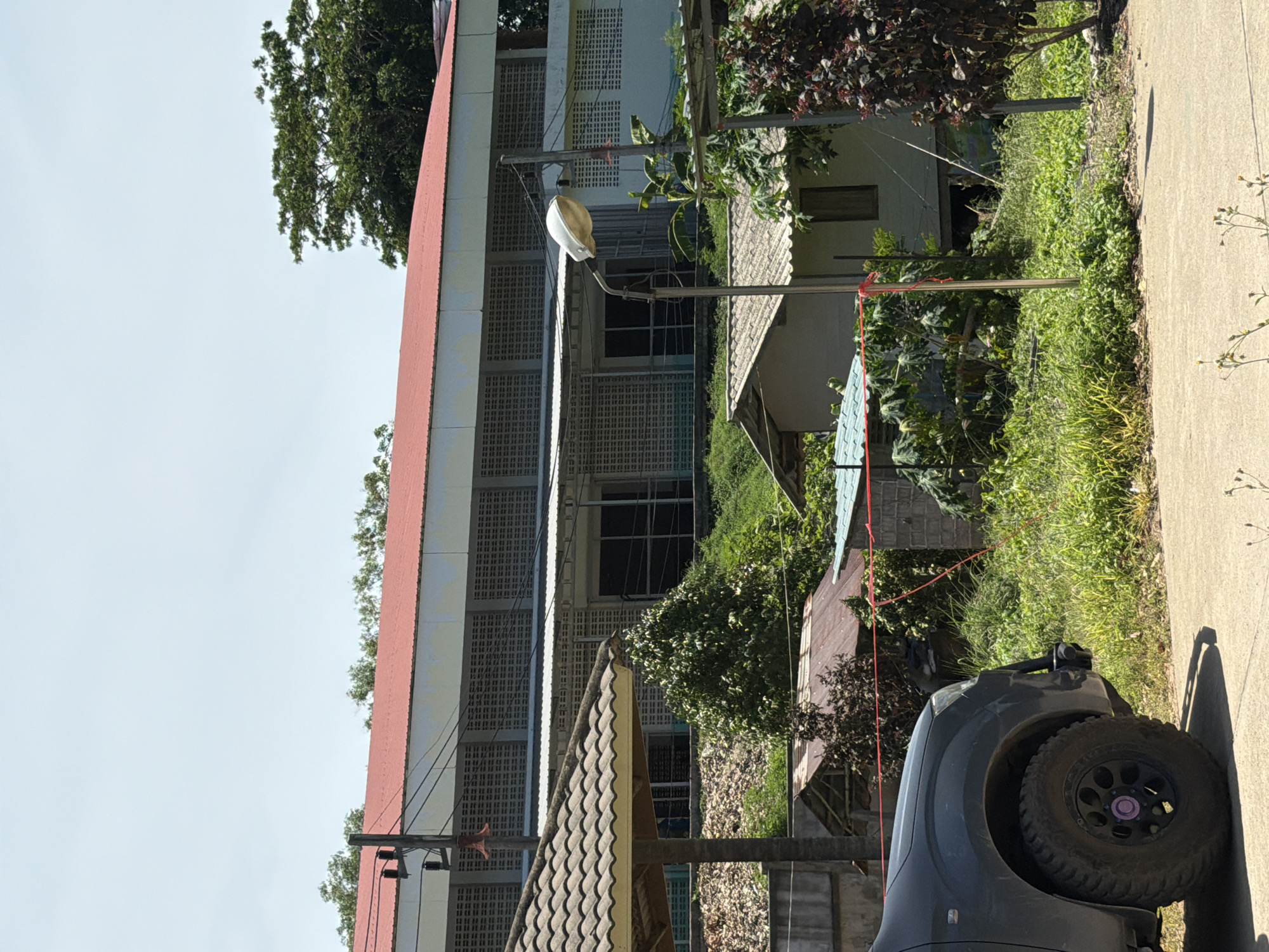 The image depicts an outdoor scene with a long, multi-story building featuring a red roof and white exterior. The building has rows of ventilation grilles. In the foreground, a dark-colored truck is partially visible on the left, with its front wheel and fender in view. A streetlamp is positioned in the middle ground, along with several smaller structures, some with corrugated metal roofs, and dense green vegetation. The ground appears to be a paved surface in the immediate foreground and rougher terrain with grass and foliage behind it. The sky is a clear light blue. The location is identified as Baan Bo Phut, Thailand.