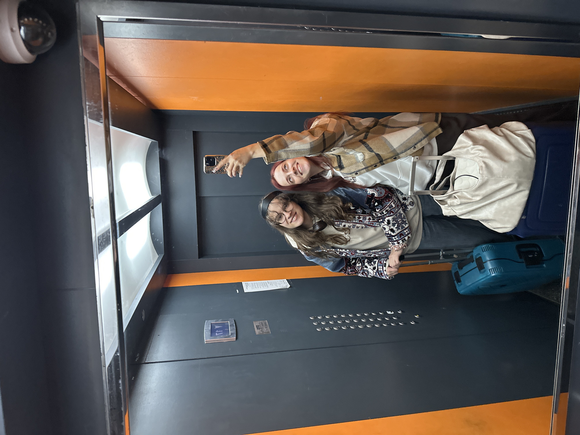 Two young women are taking a selfie in the mirror of an elevator, posing with suitcases and bags. The elevator has a modern design with gray walls and orange accents at the top and bottom. Control panels with buttons are visible on the elevator wall. A security camera is mounted near the mirror.