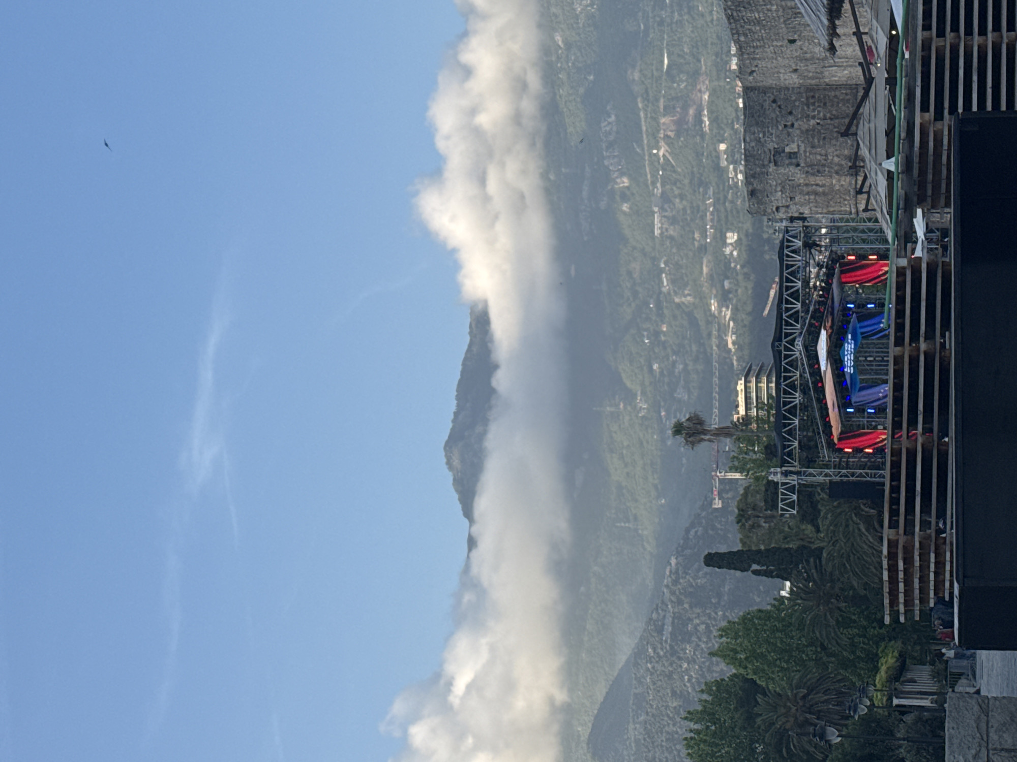 The image shows a mountainous landscape with a partial view of the city of Budva, Montenegro. A mountain range is partially obscured by low-lying clouds. In the foreground, a construction site or outdoor event area is visible, marked by scaffolding and a stage with red and blue lights. The right side of the image includes wooden structures, perhaps part of the construction or venue. The sky is pale blue with a few wispy clouds, and what appears to be a bird is flying. The location is Budva, Montenegro, based on the recognizable mountain scenery and urban elements.