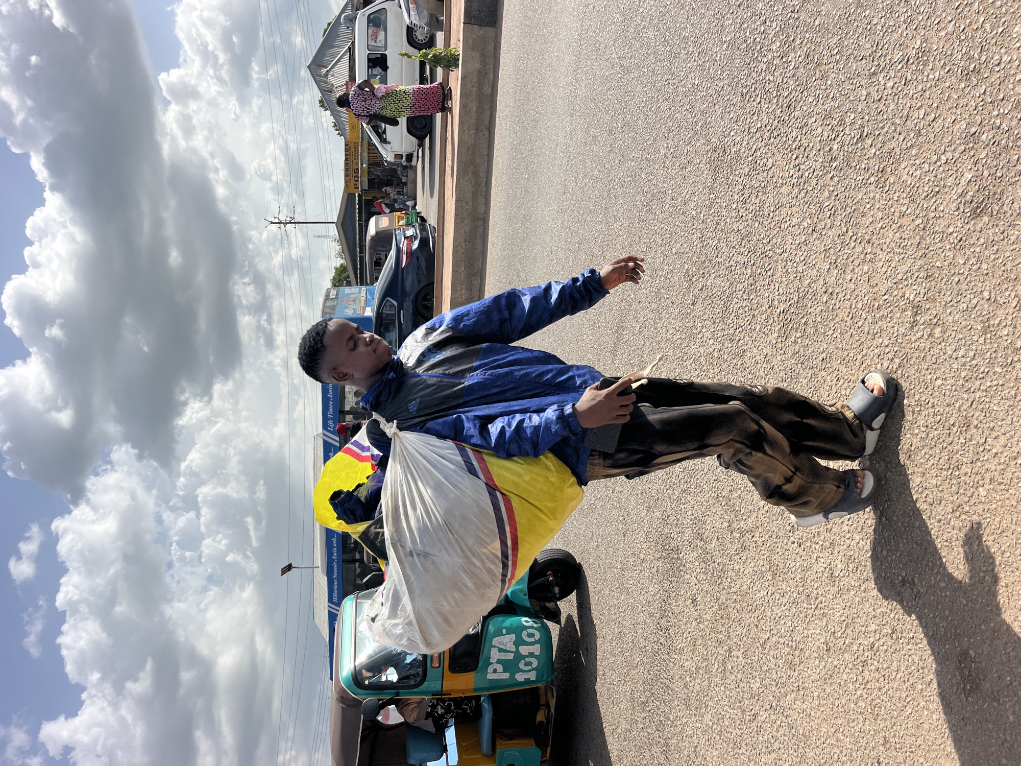 A young man, wearing a blue and black jacket, brown patterned pants, and grey sandals, stands on a paved road next to a small three-wheeled vehicle. He is carrying a large white and yellow bag slung over his shoulder. The vehicle, which appears to be a tuk-tuk or auto-rickshaw, is turquoise and yellow and has the registration number "PTA-10108" visible on its side. The man has a relaxed posture, with his left arm outstretched and his right hand holding a mobile phone.
In the background, there is a row of shops with awnings. A white van is parked in front of one of the shops, and a woman in a colorful dress is standing nearby. The sky is filled with large, fluffy white clouds, suggesting it is daytime, likely late morning or afternoon, with fair weather. The scene appears to be a roadside marketplace or a busy street in a town or city.
Visible text includes "PTA-10108" on the vehicle and on a sign behind the man, "Life Times, Amin...", and "Hilderman Sannie, Amin arch...". The overall atmosphere seems to be one of everyday life and commerce.