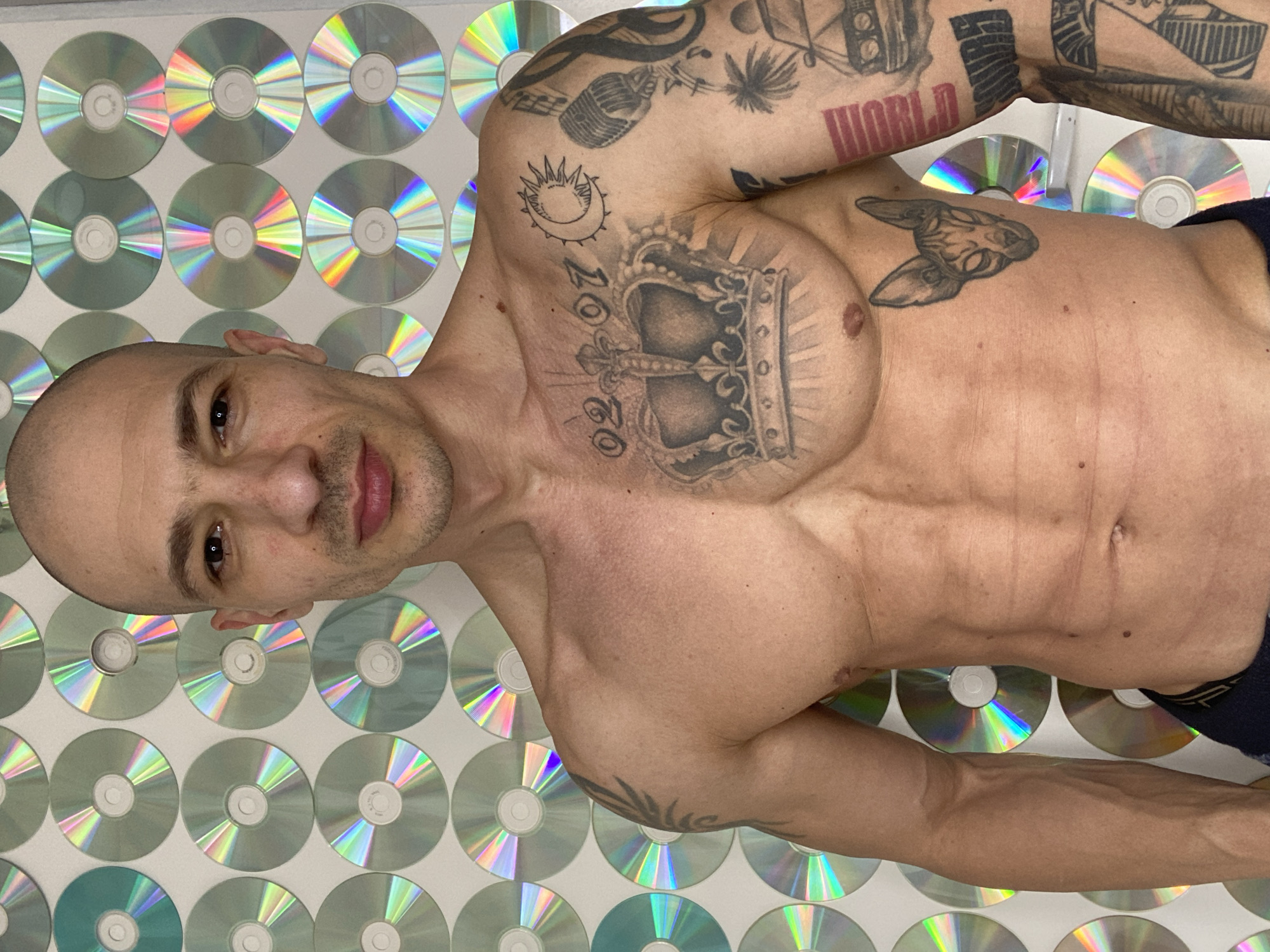 An adult male is depicted in a waist-up, frontal portrait against a wall adorned with numerous compact discs (CDs). The subject is bald, with a fair to medium complexion, and a neutral expression, gazing directly at the viewer. He is shirtless, revealing a muscular build.
Distinguishing tattoos include a large crown centered on his chest, flanked by the numbers "02" and "07." A crescent moon and stylized sun tattoo are located on his upper left chest. His left arm (viewer's right) features multiple tattoos, including a microphone, musical note, palm trees, a vintage car, and text resembling "WORLD." A Sphynx cat head tattoo is positioned on his lower right chest (viewer's left). Tribal or abstract designs are visible on his right shoulder and upper arm (viewer's left).
The background wall is entirely covered by a grid arrangement of reflective CDs, which exhibit iridescent light patterns. The waistband of dark undergarments is partially visible at the bottom of the frame. This scene is situated indoors, within the city of Pleven, Bulgaria.