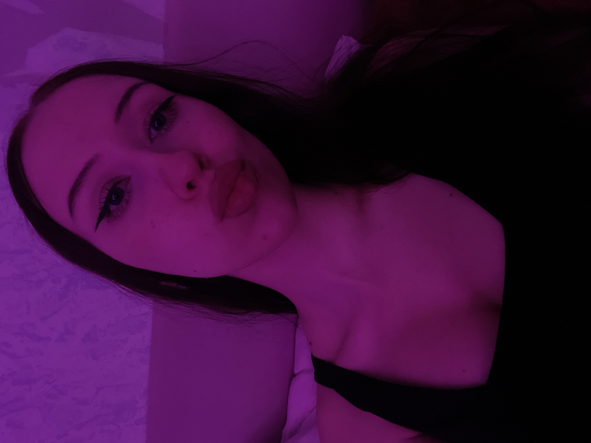 This is a medium-shot selfie of a young woman with dark hair, taken indoors under purple lighting. She is wearing a black tank top. Her head is tilted slightly, and she is looking towards the camera with her lips pursed. The background appears to be a textured wall and a bed. There is no indication of the specific location within Votkinsk, Russia.