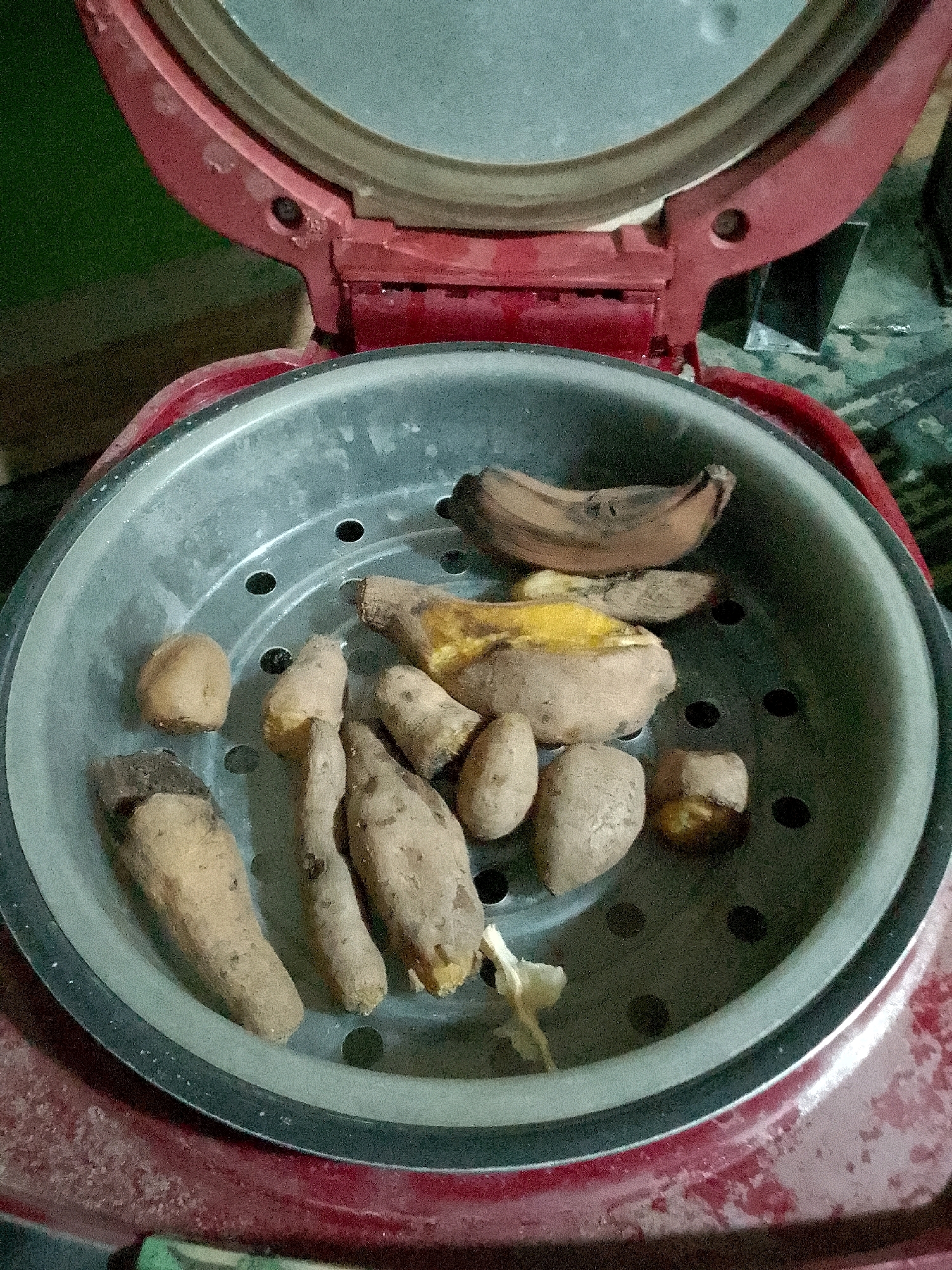 The image shows a red rice cooker with its lid open, revealing a metal steaming basket filled with food items. The steaming basket contains several pieces of what appear to be sweet potatoes, some of which are whole and others cut in half, exposing a yellow interior. There is also one overripe banana with brown spots. The food items are arranged on the perforated surface of the steaming basket. The exterior of the rice cooker is red, and the inner pot is silver. The background is blurry, but a green surface is visible to the left, and a dark, cluttered area is to the right. There are no people visible in the image, and no specific location information is provided within the image itself to confirm it is in Gunungmasigit, Indonesia. The scene is indoors, likely in a kitchen setting.