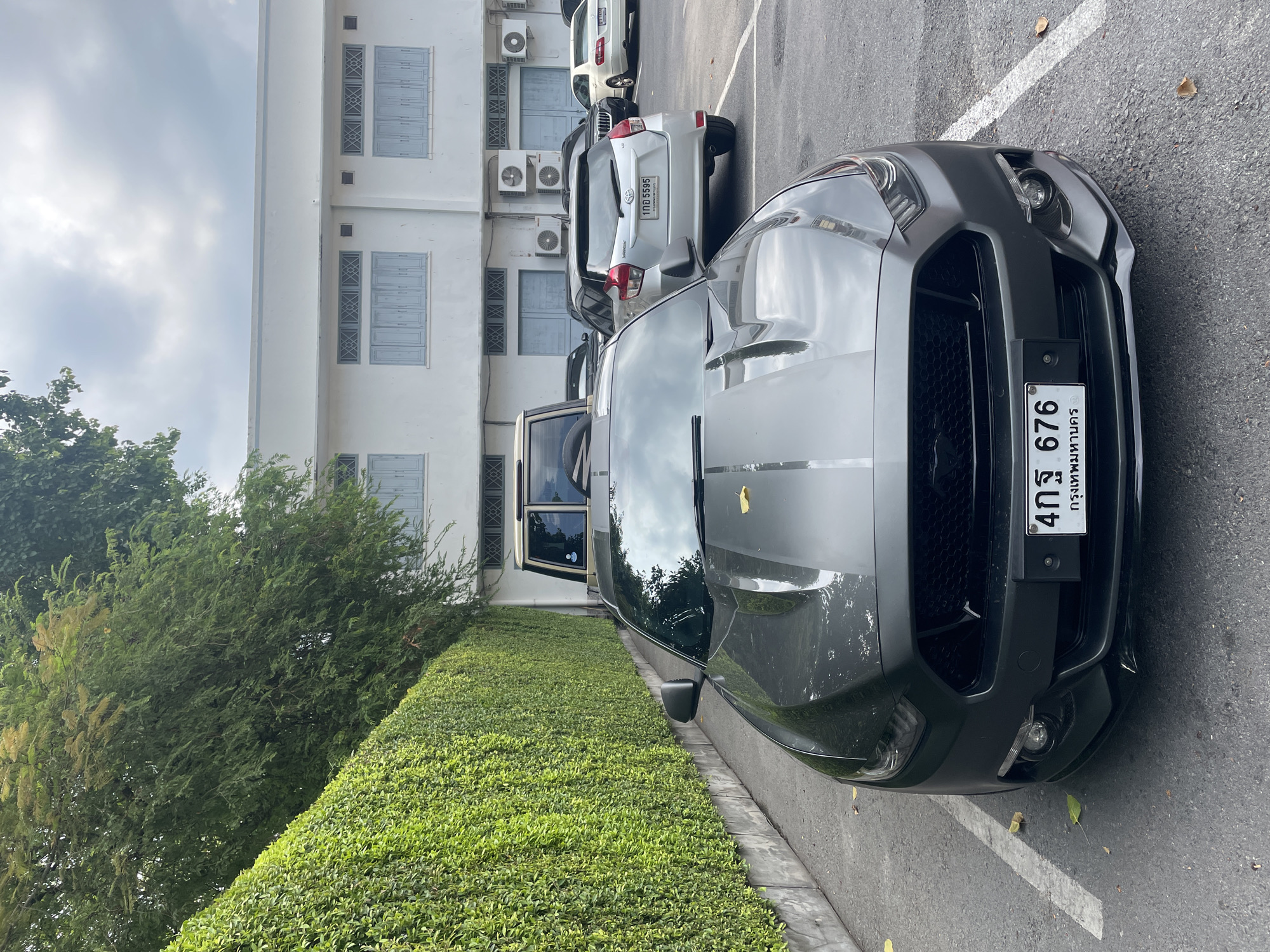 A gray Ford Mustang is parked in a parking lot in Bangkok, Thailand. The license plate reads "4กร 676." Behind the Mustang are other parked vehicles and a white building with light blue trimmed windows and several air conditioning units. A green hedge separates the car from the building. The sky is overcast.