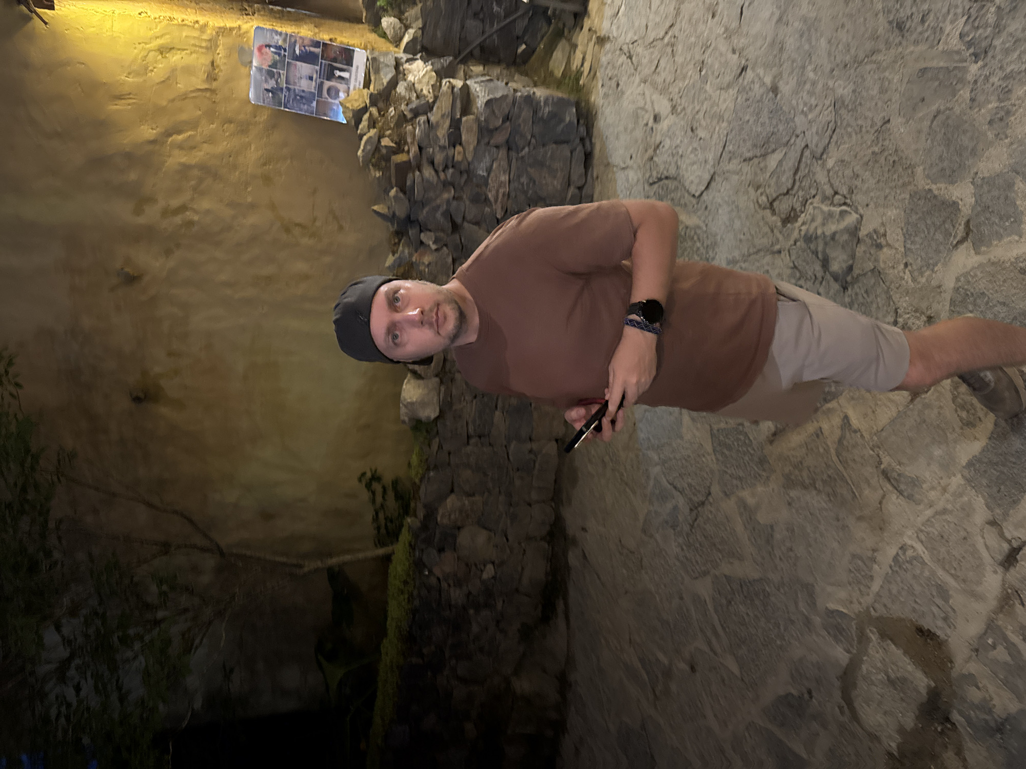 The image shows a man in Lũng Cú Commune, Vietnam. He is wearing a brown t-shirt, khaki shorts, and a black cap. He is holding a black smartphone in his right hand. The background includes a stone wall and a building. A photo collage hangs on the wall. The ground is paved with stone. The lighting suggests it is evening or night. The wall has a yellow tone to it. Some plants are growing on the wall near the ground.