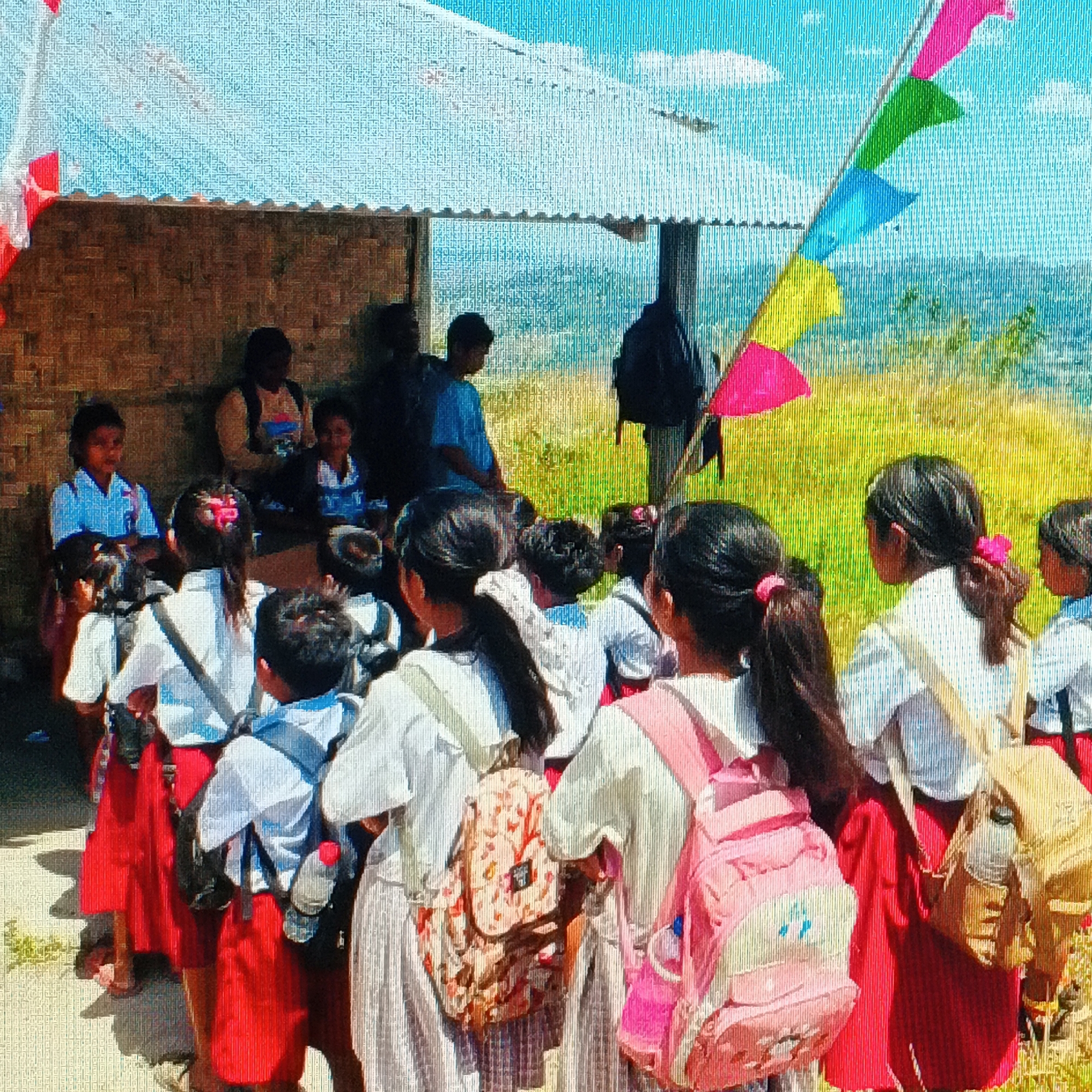 In Ende, Indonesia, a group of children, likely students, are gathered outside a simple open-air structure with a corrugated metal roof. Most of the children are wearing school uniforms, consisting of white shirts and red skirts or shorts. Some are carrying backpacks. The setting appears to be a rural or semi-rural area, with a grassy hillside in the background. Colorful flags are strung on a pole on the right side of the image, and red and white flags are visible on the left. The overall atmosphere suggests a school gathering or event.