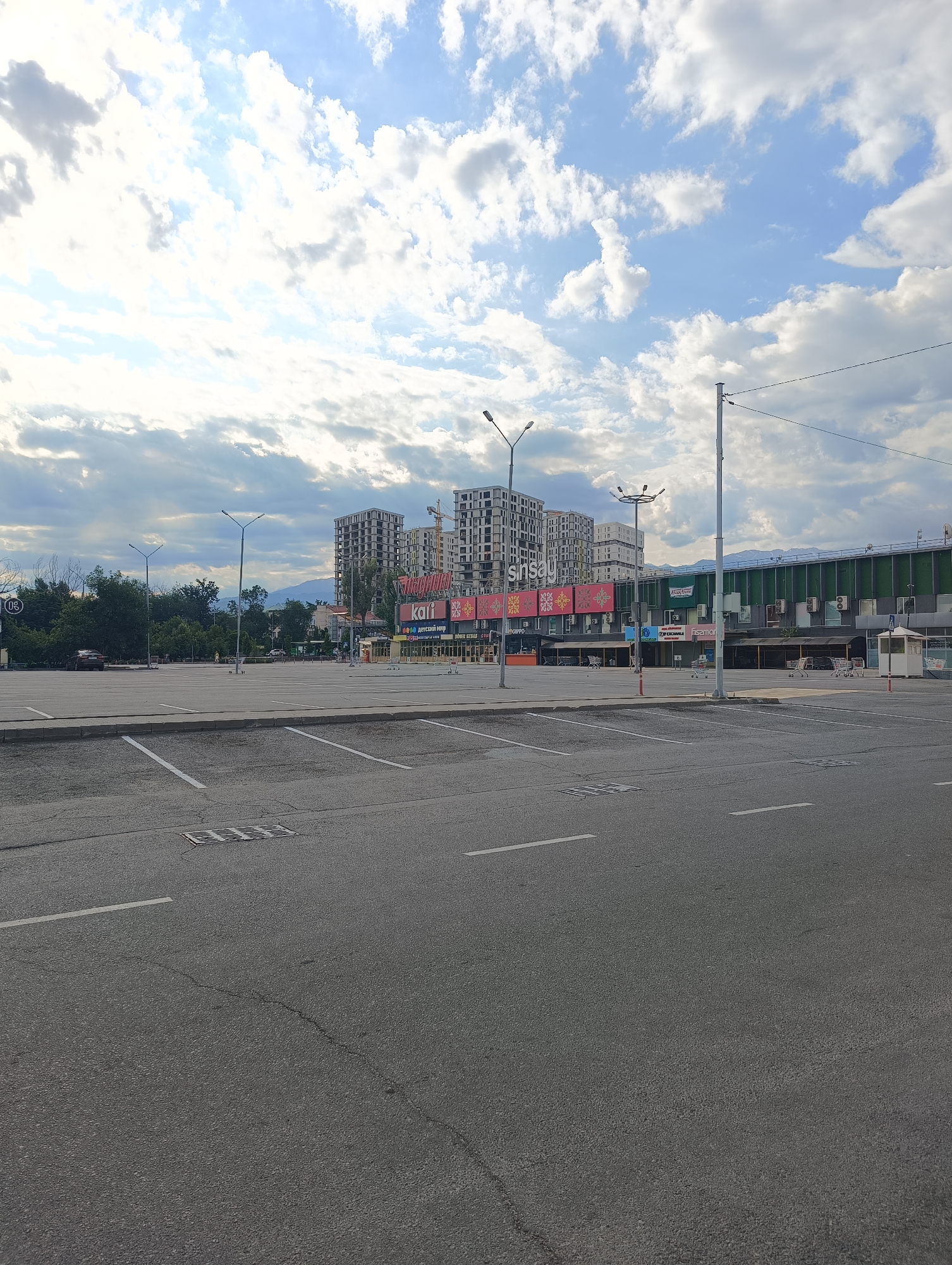 This is a street-level, eye-level, wide shot of an empty parking lot in front of a commercial building in Almaty, Kazakhstan. The sky is overcast with large, fluffy white clouds. The parking lot is paved and marked with white parking lines. There are several streetlights and utility poles scattered throughout the lot. In the background, there is a large, modern commercial building with a green facade and several windows. Above the commercial building, there are several tall, unfinished residential buildings under construction, with a crane visible on one of them. Some of the commercial building's signage is visible, including "snsau" and "kai." There are a few shopping carts in the parking lot and a small security booth near the right side of the commercial building. In the distance, mountains can be seen. The overall scene suggests a commercial area with ongoing construction.