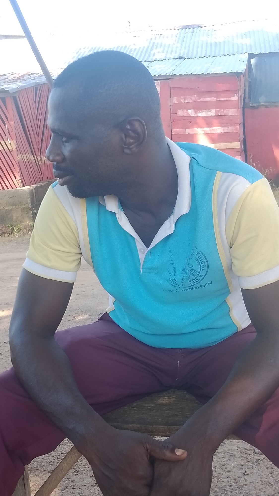 A man with dark skin and a close-cropped haircut is sitting on a wooden bench outdoors. He is wearing a collared polo shirt with light blue and yellow panels, and maroon pants. The man is looking to his left, with his arms resting on his lap and his hands clasped. The background shows a corrugated metal roof over a red wooden structure, possibly a building. The ground appears to be dirt. The scene is located in Adankolo, Nigeria.