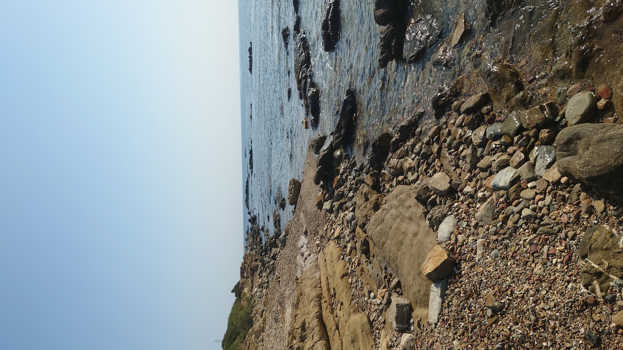 The image depicts a rocky coastline under a clear blue sky. The foreground is dominated by a shore composed of various sizes of stones and rocks, with some larger flat rock formations present. The water's edge is visible, with gentle waves lapping against the rocks and a few submerged rocks. The sea extends to the horizon, meeting the sky in a clear line. To the left, a grassy slope with some vegetation leads up to a higher vantage point, with a distant, indistinct object that might be a utility pole. The overall scene suggests a natural, undeveloped coastal area. The location in Biddeputzi/Villaputzu, Italy, is not visually evident from the image itself.
