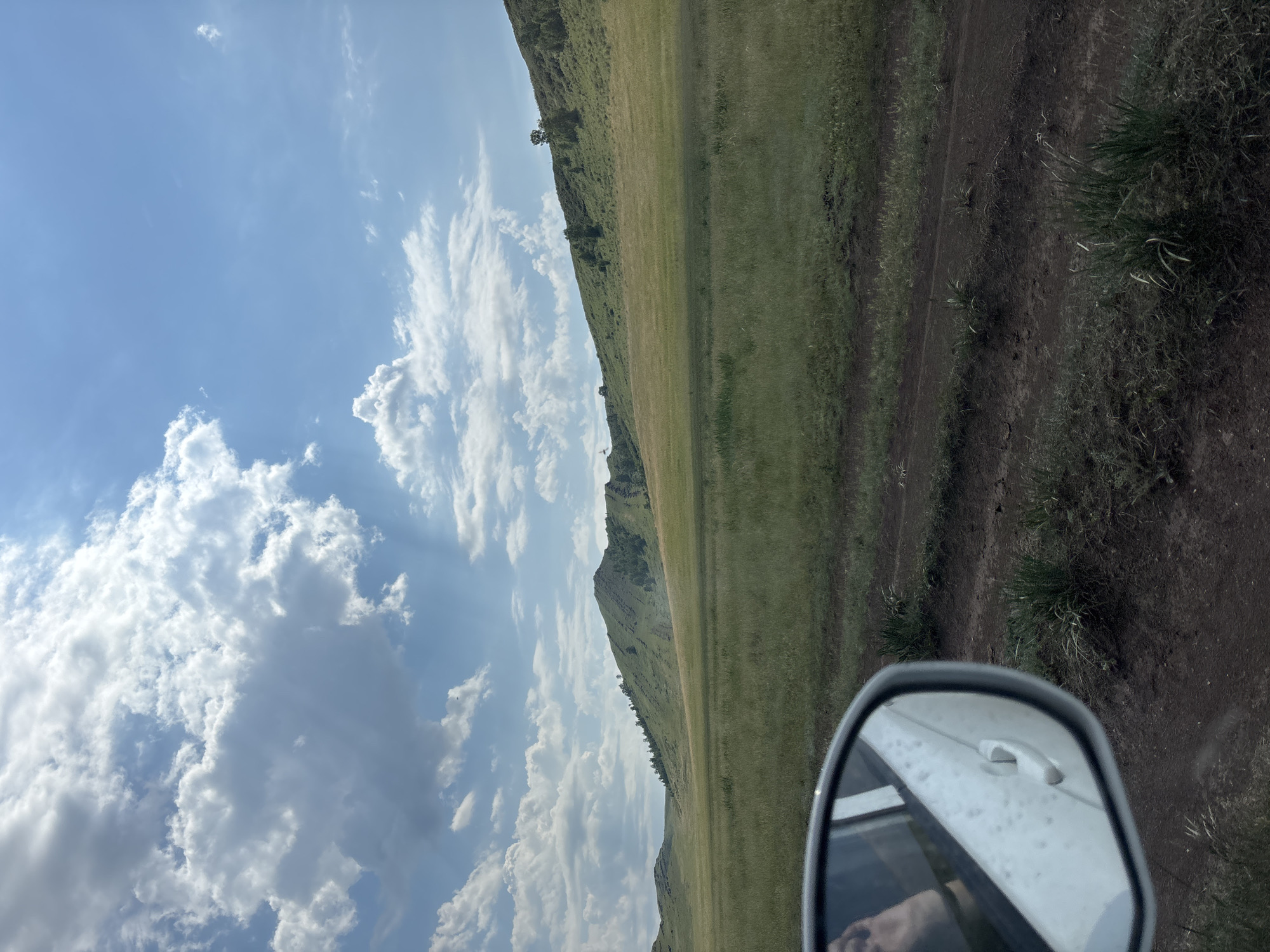 The image captures a rural landscape viewed from inside a vehicle, with a side mirror prominently featured in the lower left quadrant reflecting a person's hand and arm. The scene depicts a vast, grassy plain stretching towards rolling, green hills under a bright blue sky dotted with fluffy white clouds. A dirt road is visible in the foreground, winding through the sparse vegetation. The overall impression is one of a serene, open natural environment, possibly in the outskirts of Троицкое, Russia, given the provided context.