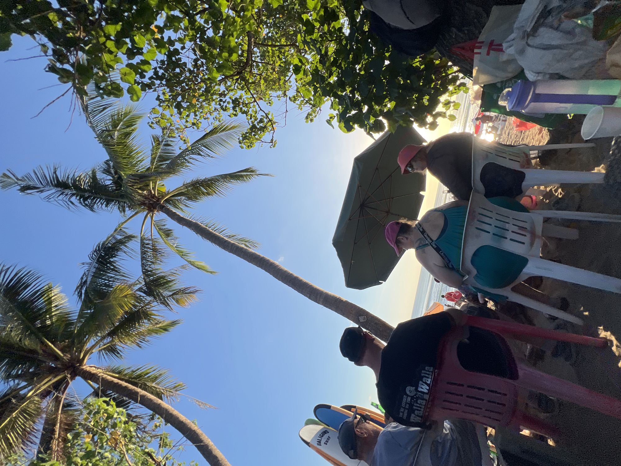In Kuta, Indonesia, a group of people are seated on chairs on a beach, facing the ocean. Palm trees and lush green foliage frame the scene, with the bright sun casting long shadows across the sand. Several surfboards are propped up nearby, suggesting a surf-friendly location. In the foreground, three individuals are visible from behind, wearing casual attire. One person is wearing a black t-shirt with text that reads "Walla Walla". Another person, wearing a green tank top and a pink hat, is seated next to them. A third person, wearing a dark long-sleeved shirt and a pink hat, is seated to the right, looking out towards the ocean. In the background, other beachgoers can be seen enjoying the sun and surf. The overall atmosphere is relaxed and tropical, indicative of a beach resort or popular tourist destination.