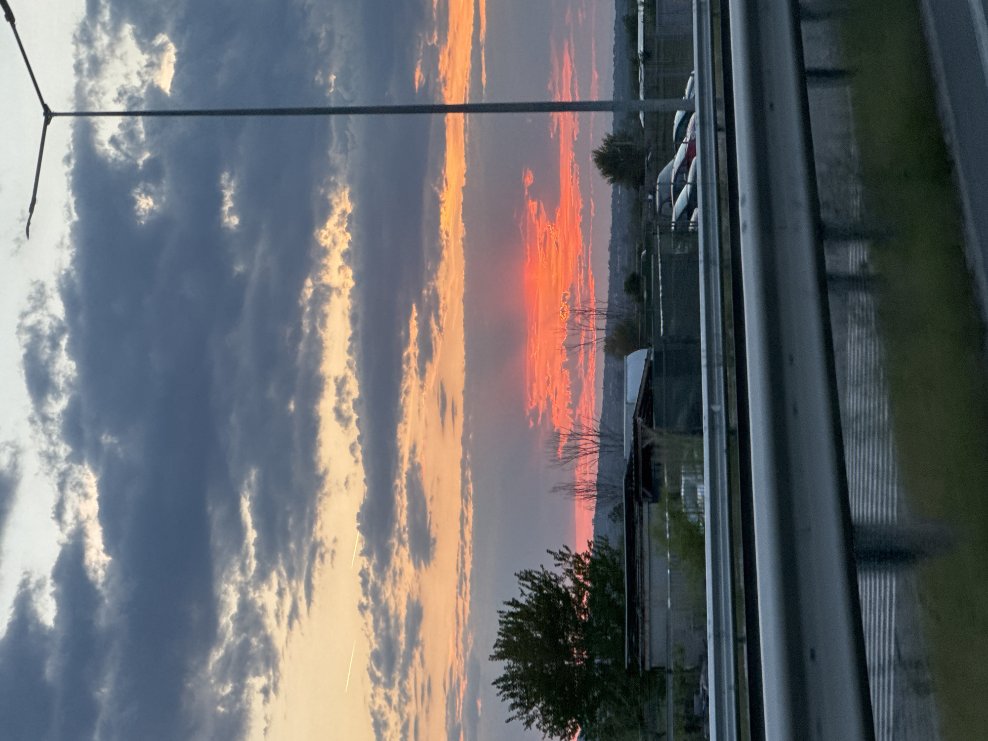 The image captures a vibrant sunset from what appears to be a roadside perspective, possibly near or along a highway. In the foreground, a metal guardrail runs along the right edge, partially obscuring the view. A light pole stands on the left, its arm extending into the frame. The sky is filled with dramatic clouds, ranging from dark gray to orange and red hues illuminated by the setting sun. The horizon is somewhat obscured by trees and buildings, suggesting a suburban or industrial area. A few cars are visible parked in the distance. It is not possible to confidently ascertain the specific location in Asenovgrad, Bulgaria from the image alone.