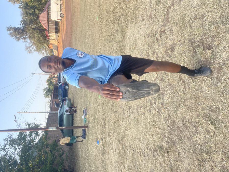 A man in a light blue shirt and black shorts stretches his leg. He is holding the sole of his shoe with his hand. He wears black socks and the field is a dry grass. In the background, there are cars parked, a metal pole, and a few people sitting near the cars. A building with a trailer is further in the background, under a clear sky.