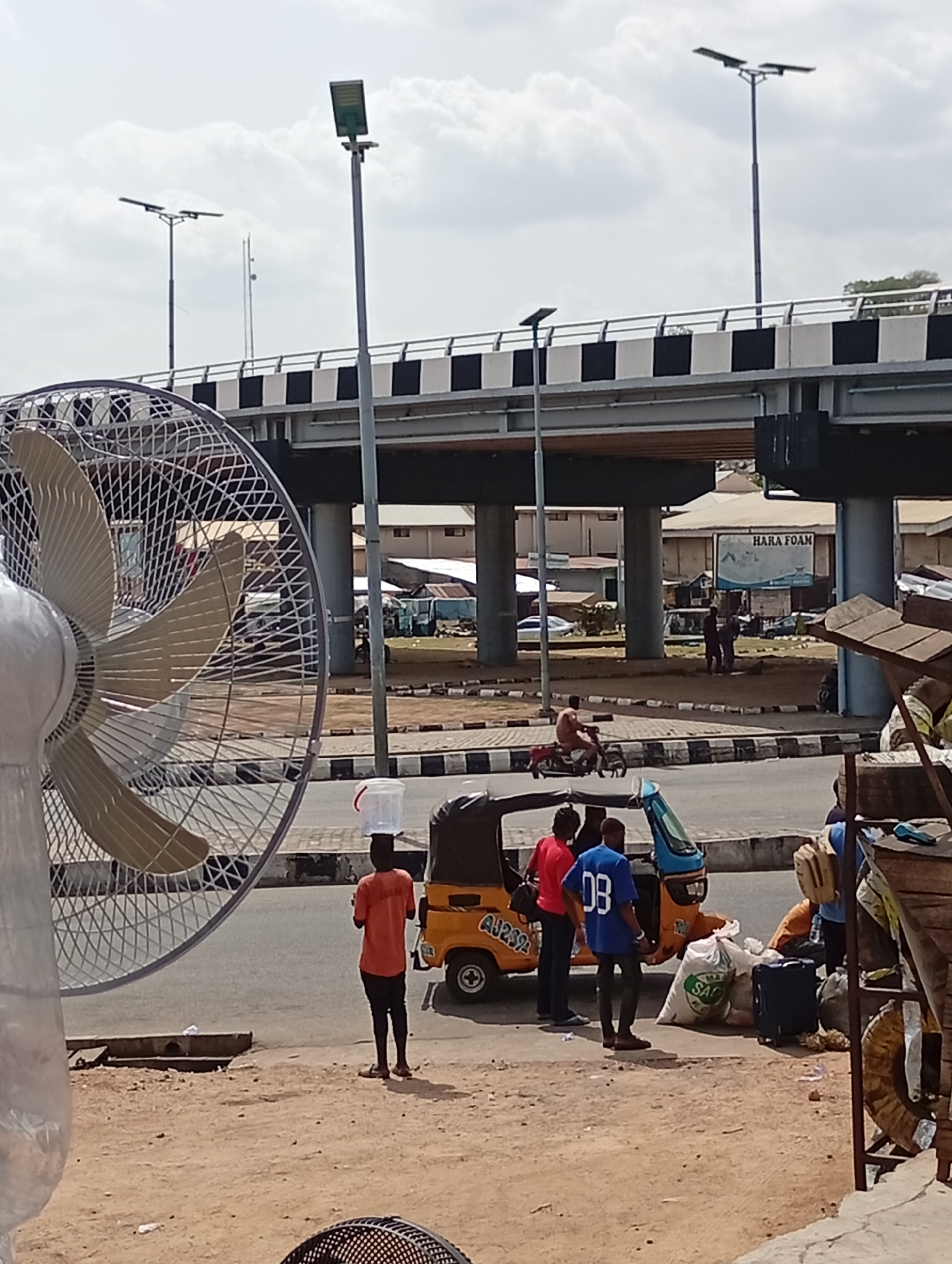 The image shows an outdoor scene in Lokoja, Nigeria, featuring a busy street and a raised highway overpass. Several people are visible near a yellow and blue "Keke Napep" tricycle with the registration number AU239. One individual is carrying a bucket on their head. A fan is positioned prominently in the foreground, partially obscuring the view. The raised highway features black and white checkered barriers and streetlights. Buildings and a "HARA FOAM" billboard are visible in the background. A motorcyclist is riding in the middle distance. Suitcases and bags are on the ground near the tricycle, suggesting a point of transport or commerce.