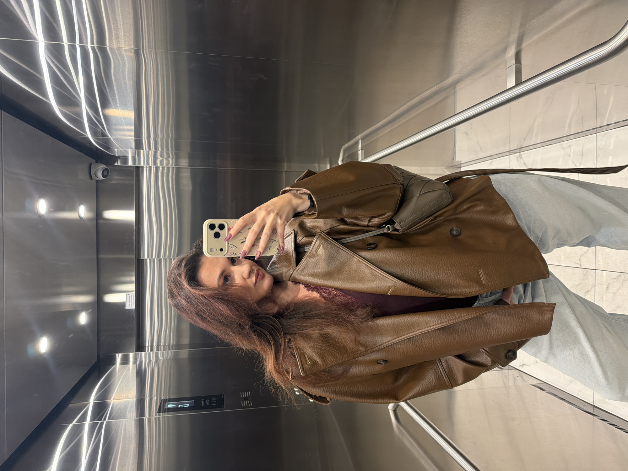 A woman with long, auburn hair stands in a modern elevator with brushed metal walls and a tiled floor. She is wearing a brown leather jacket over a maroon top and light-colored pants or jeans. She holds a cell phone with a floral design on the case in her right hand, seemingly taking a selfie in the elevator's reflective surfaces. A security camera is visible in the upper-left corner of the elevator. The scene likely takes place indoors, possibly in a building or shopping mall in Belgrade, Serbia.