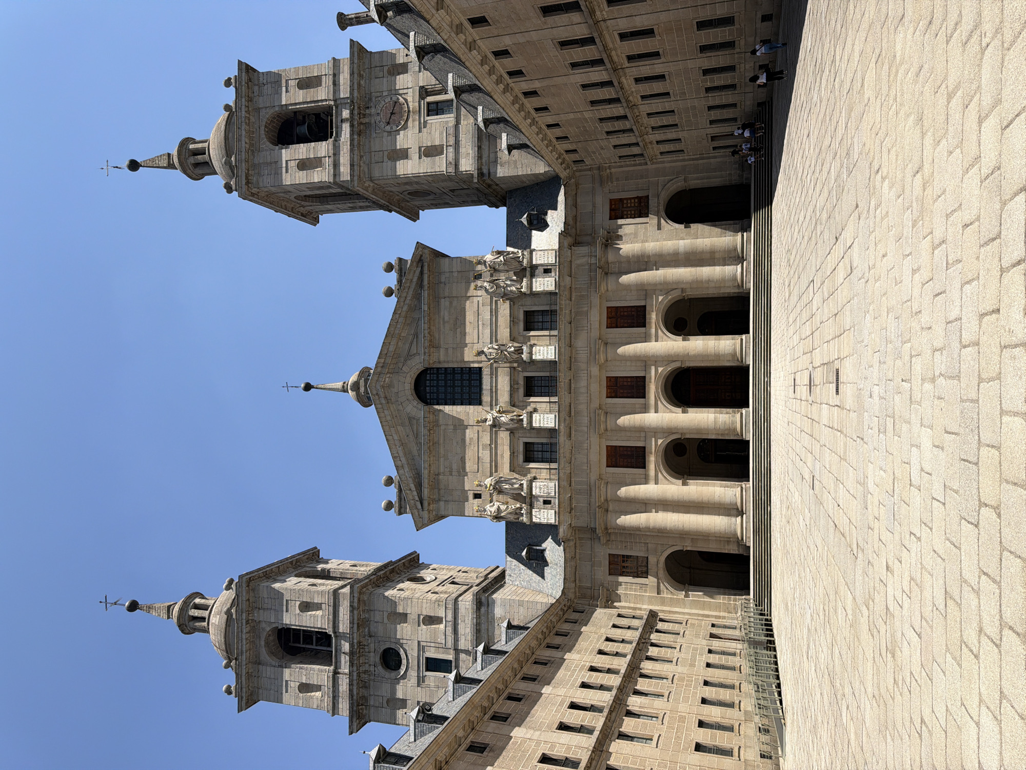 This is a vertically oriented, eye-level shot of the San Lorenzo De El Escorial in Spain. The large stone building dominates the frame with its classical architecture, featuring multiple towers, arched windows, and statues adorning the facade. The sky is a clear, bright blue, indicating a sunny day. The paving stones in the foreground are light beige, suggesting a courtyard or plaza. A few people can be seen in the distance, scattered across the plaza, observing the grandeur of the building. Their presence adds a sense of scale and human activity to the vast architectural structure. The sunlight casts shadows, highlighting the architectural details and textures of the stone. The overall mood is serene and awe-inspiring, emphasizing the historical and monumental nature of the location.