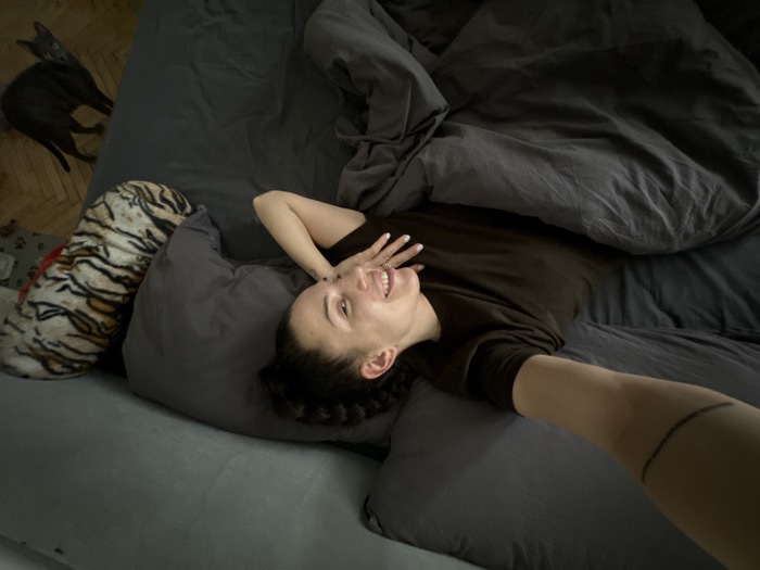 A female subject is depicted lying on a dark grey bed or large couch in an indoor residential setting. She is positioned on her back, with her head resting on a grey pillow, facing slightly upwards and towards the camera with an open-mouthed smile or laugh. Her dark hair is styled in braids or cornrows. Her left hand is raised, with white-painted fingernails visible near her face. Her right arm is extended, holding a camera or smartphone to capture a selfie; a dark, curved line tattoo is visible on her right forearm. She is wearing a dark, likely olive-green, short-sleeved top. A dark grey duvet or blanket is draped over her lower body and the bed. To her left, a long, cylindrical pillow featuring a tiger-stripe pattern is positioned against the head of the bed. In the upper left background, on a wooden parquet floor, a slender, dark-colored cat with prominent ears is visible, looking towards the female subject. A small, patterned pet mat is partially visible on the floor near the tiger pillow. The precise location within the city of Moscow, Russia, cannot be determined from the visual information provided.